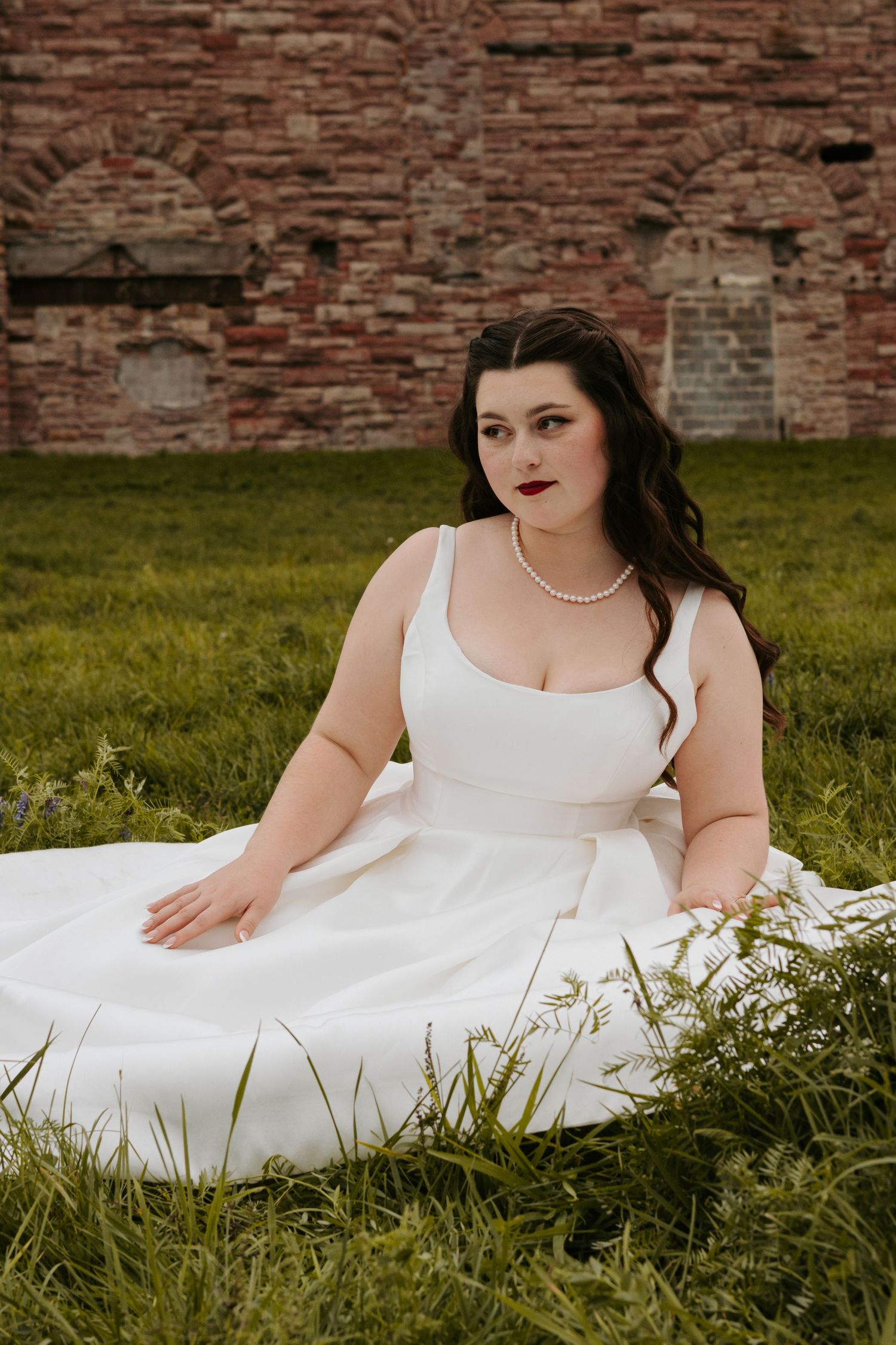 A bride in a white gown reclines gracefully on grass near a brick wall.