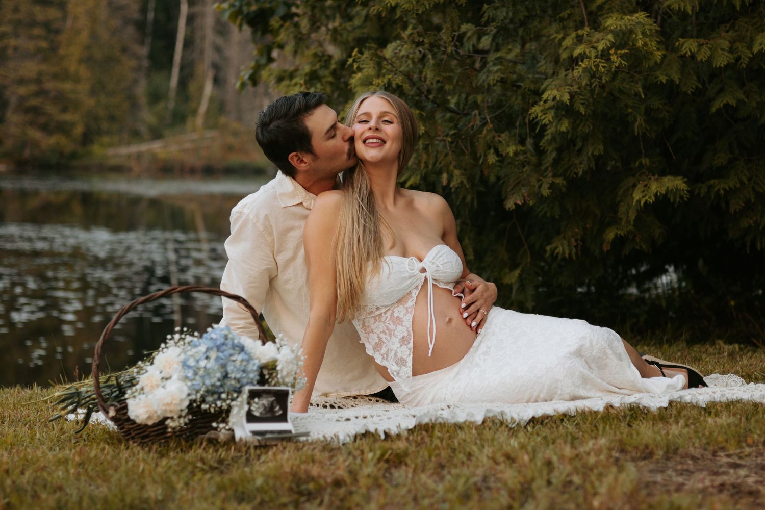 Maternity photo session by lake with couple sharing intimate moments on blanket.