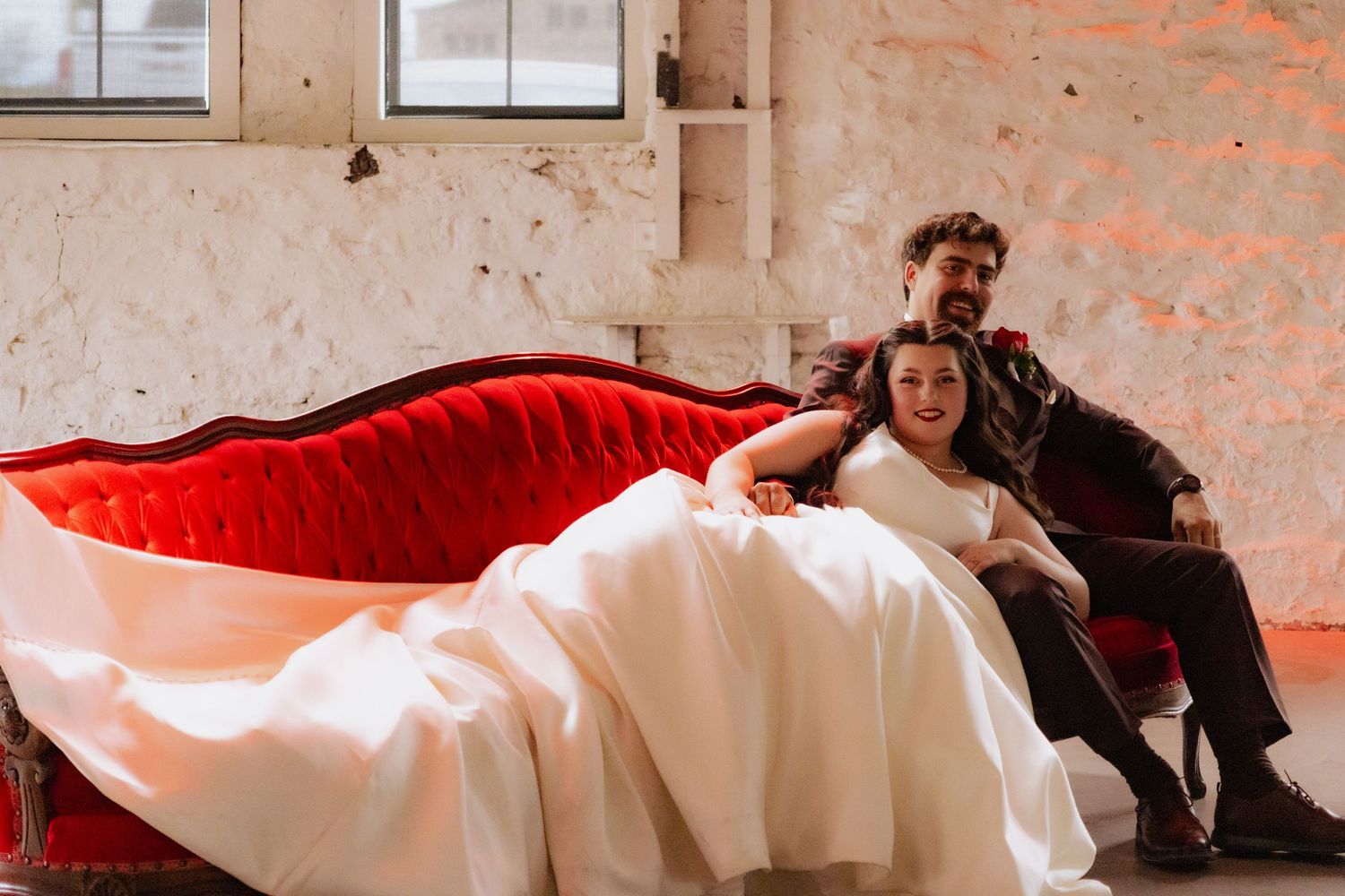Newlyweds share an intimate moment on a red velvet couch in a rustic brick room.