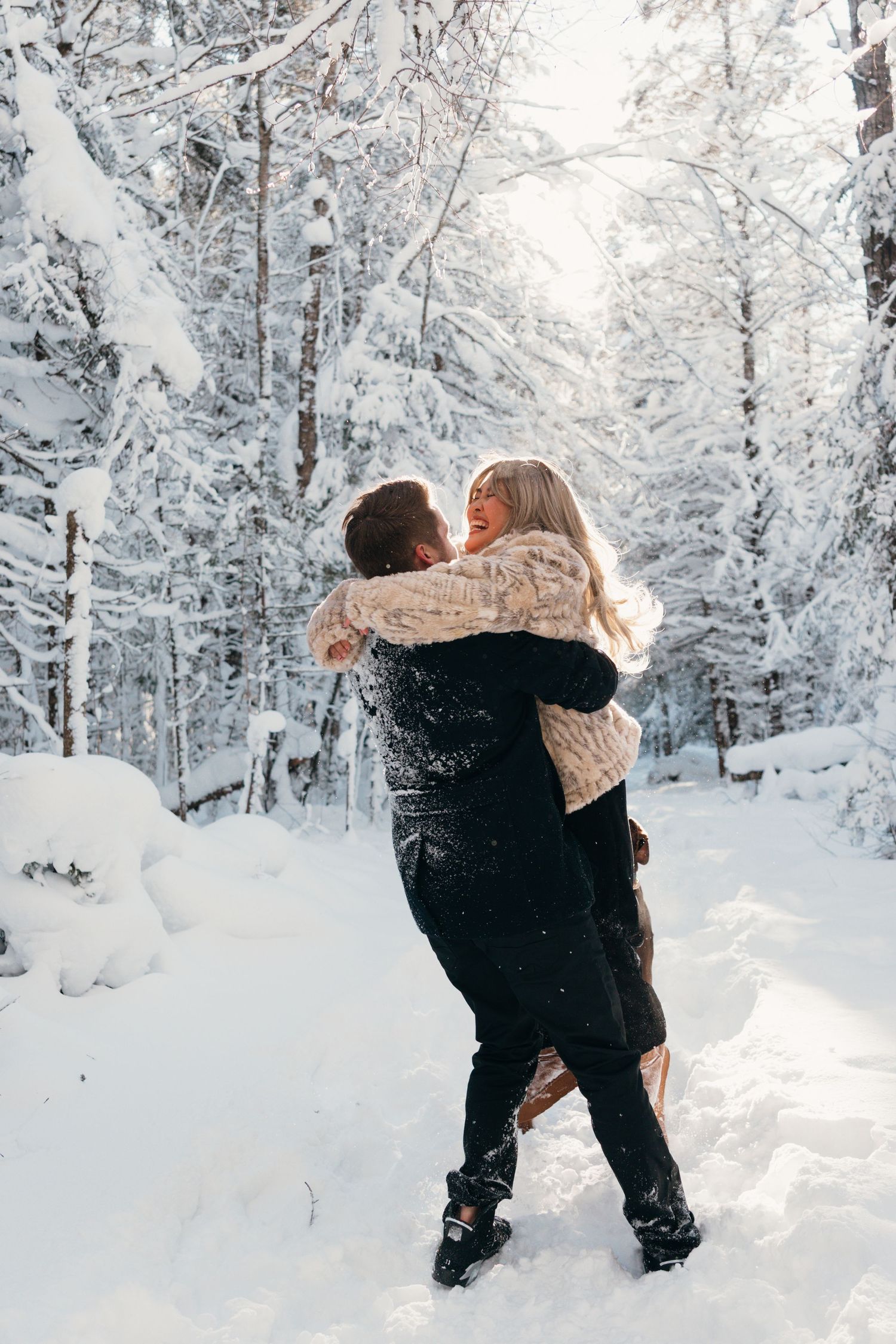 Two people embrace while playing in deep snow surrounded by snow-covered trees.