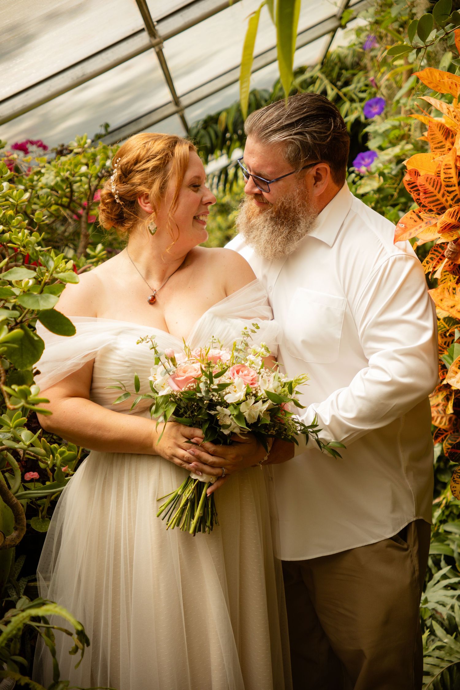 A romantic greenhouse wedding photo surrounded by colorful flowers and greenery.
