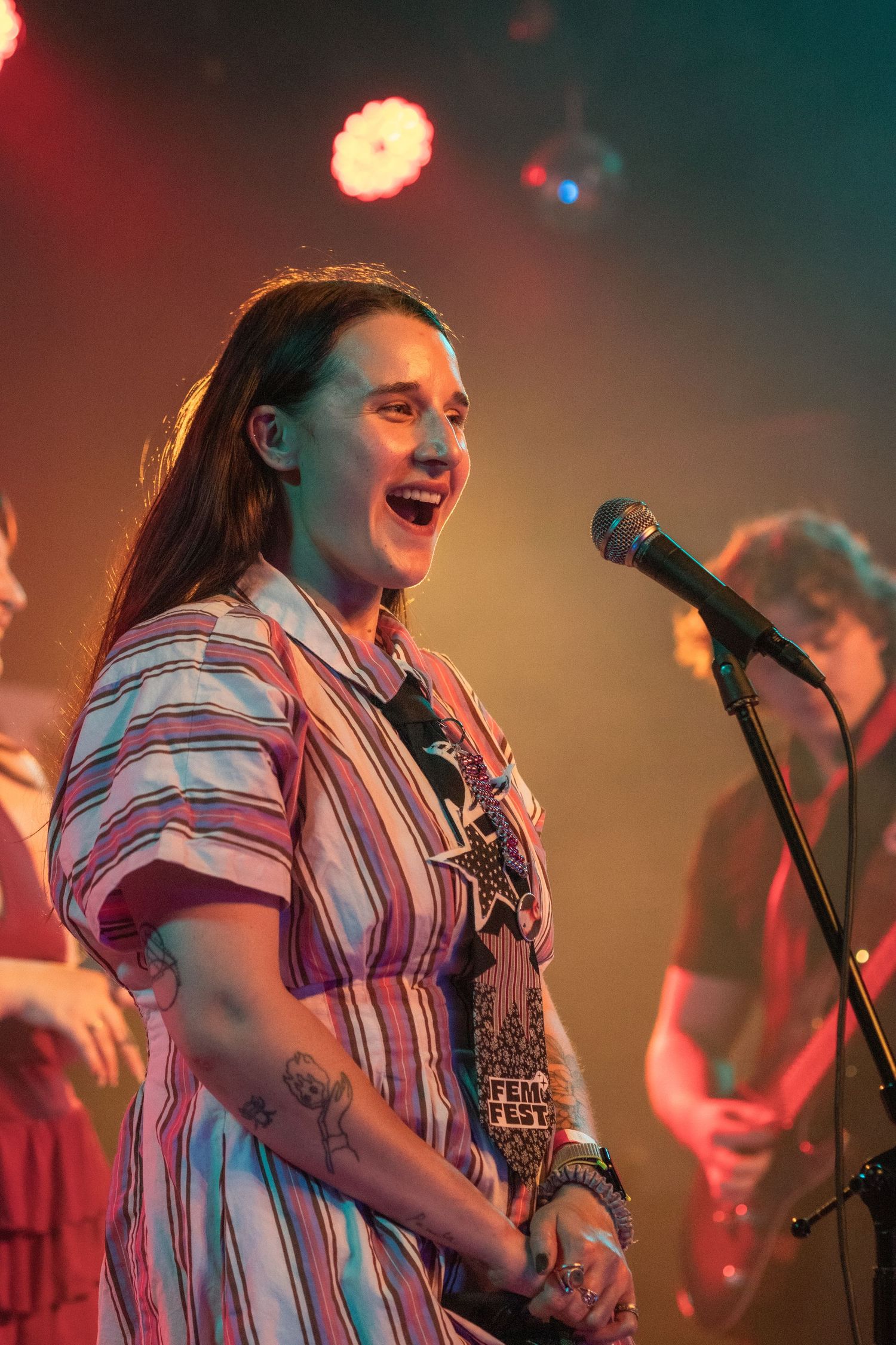 Young woman with long hair singing joyfully at a microphone on a colorful concert stage.