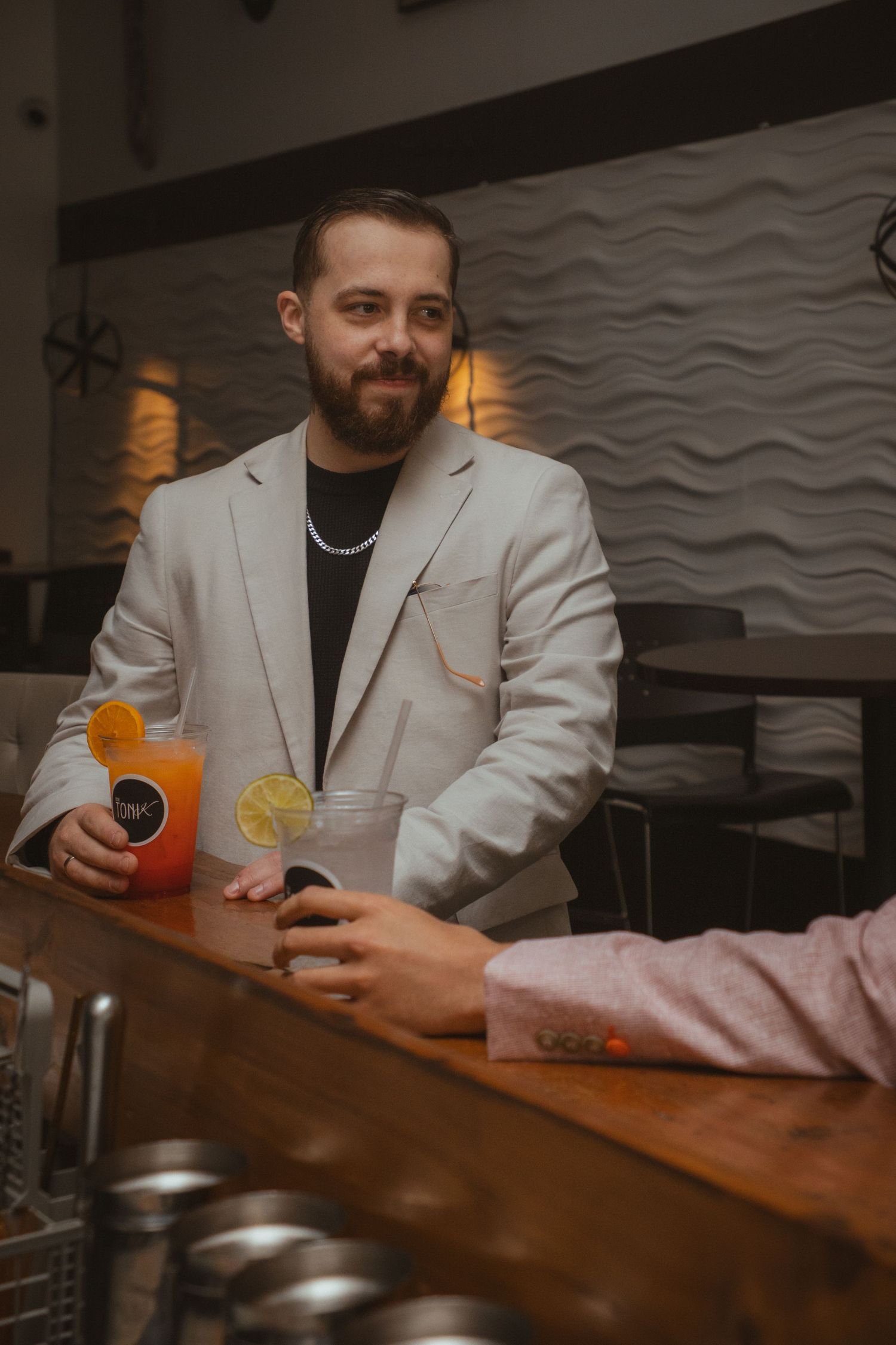 A person in a white blazer and black shirt sits at a bar with an orange cocktail.