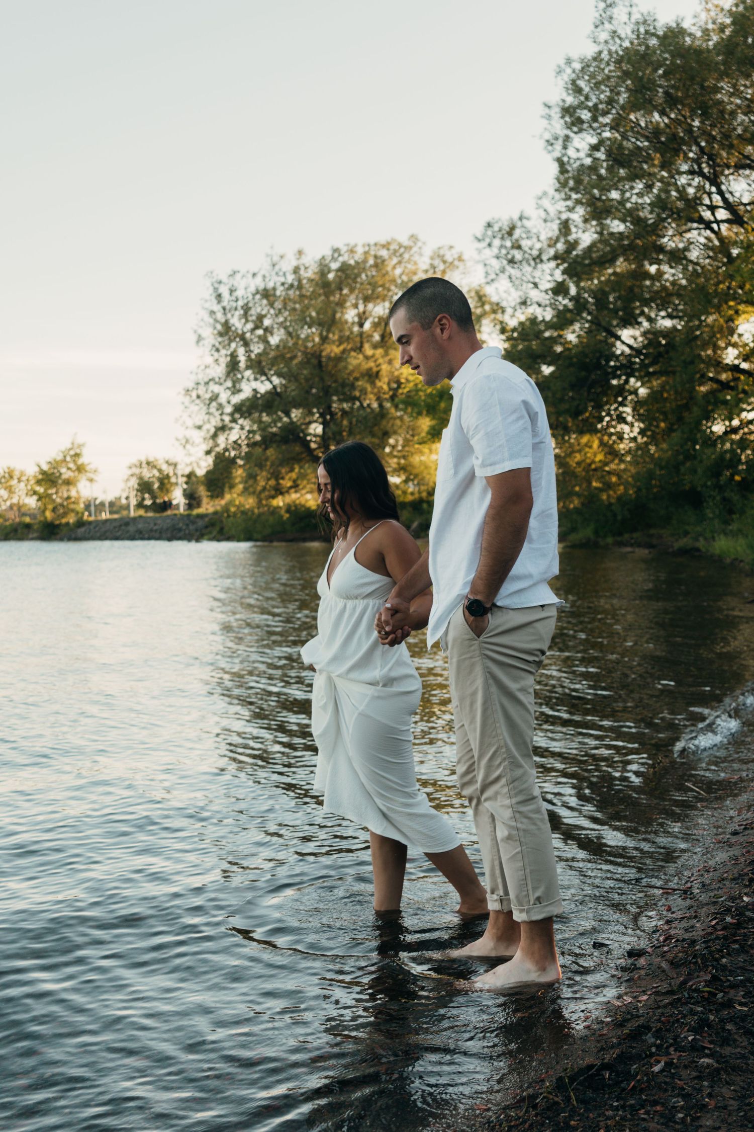 A couple in white and khaki outfits stands by the water's edge at sunset.