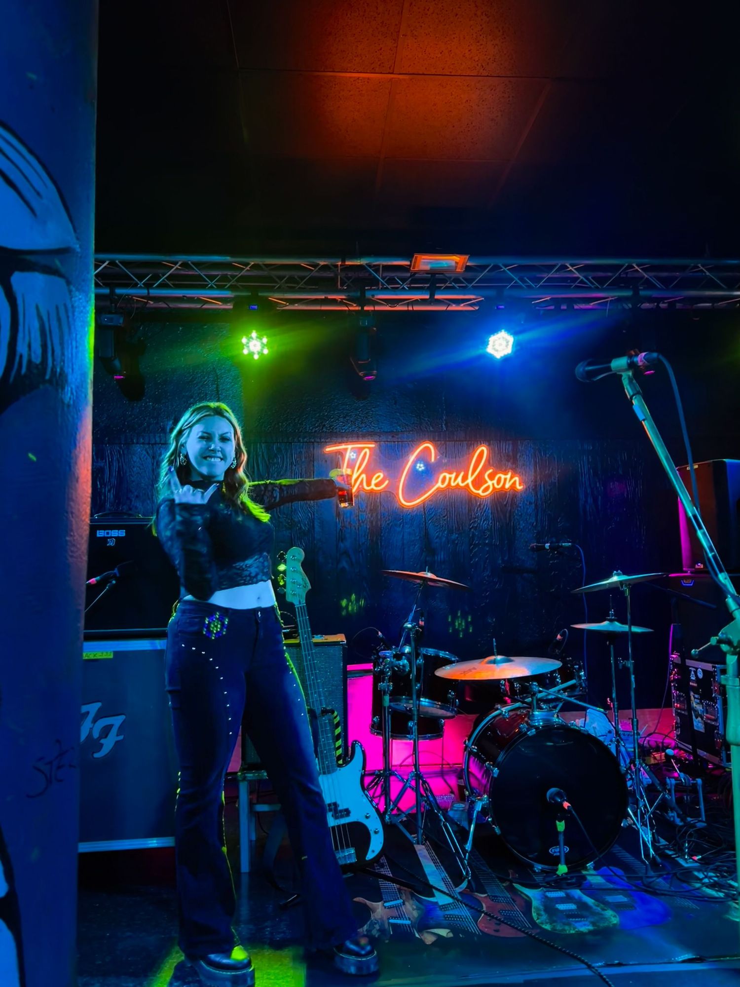 Female singer performing on stage at The Coulson venue under colorful neon lights with drum kit in background.