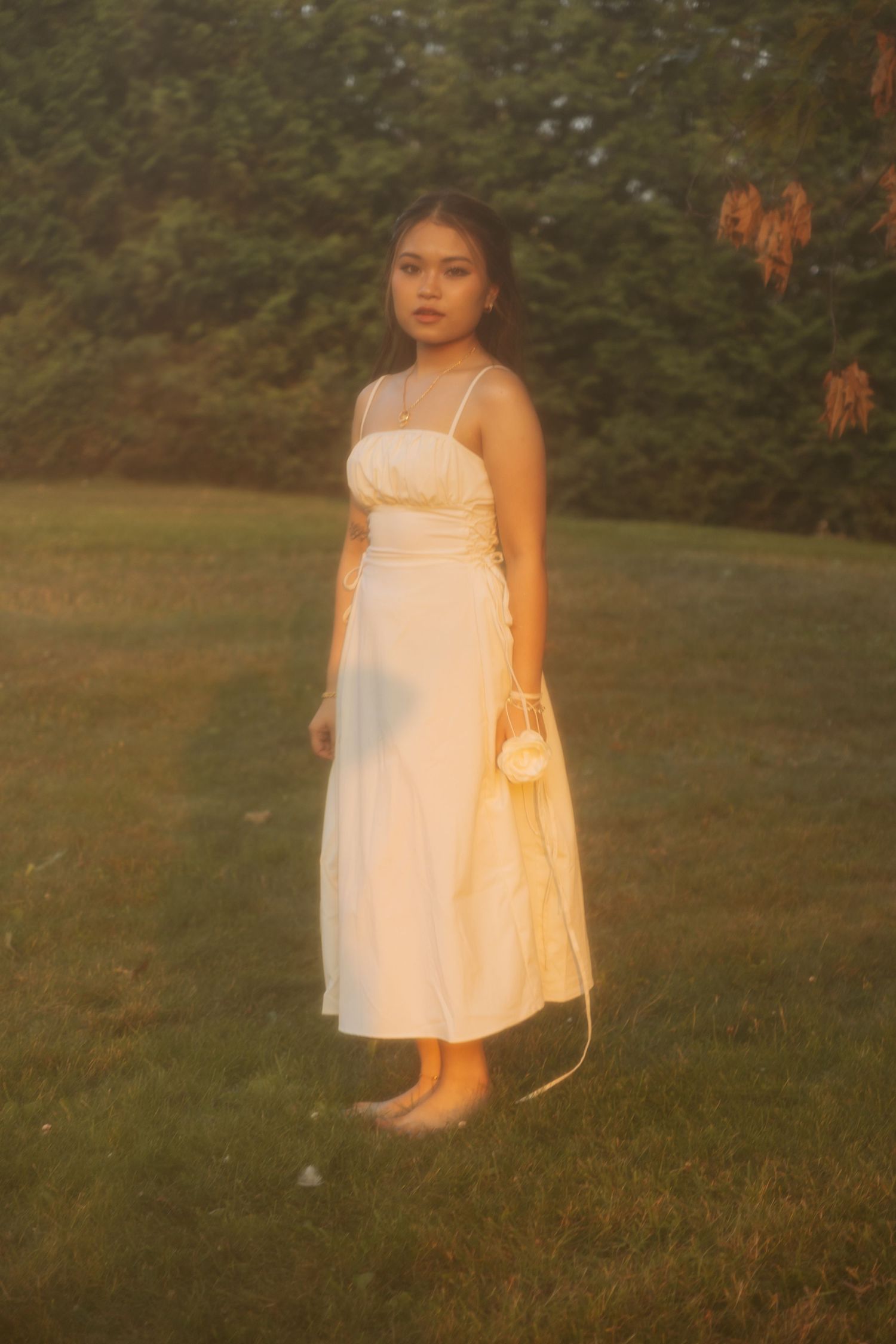 A full-length portrait showing someone in a white dress standing barefoot on grass at golden hour.