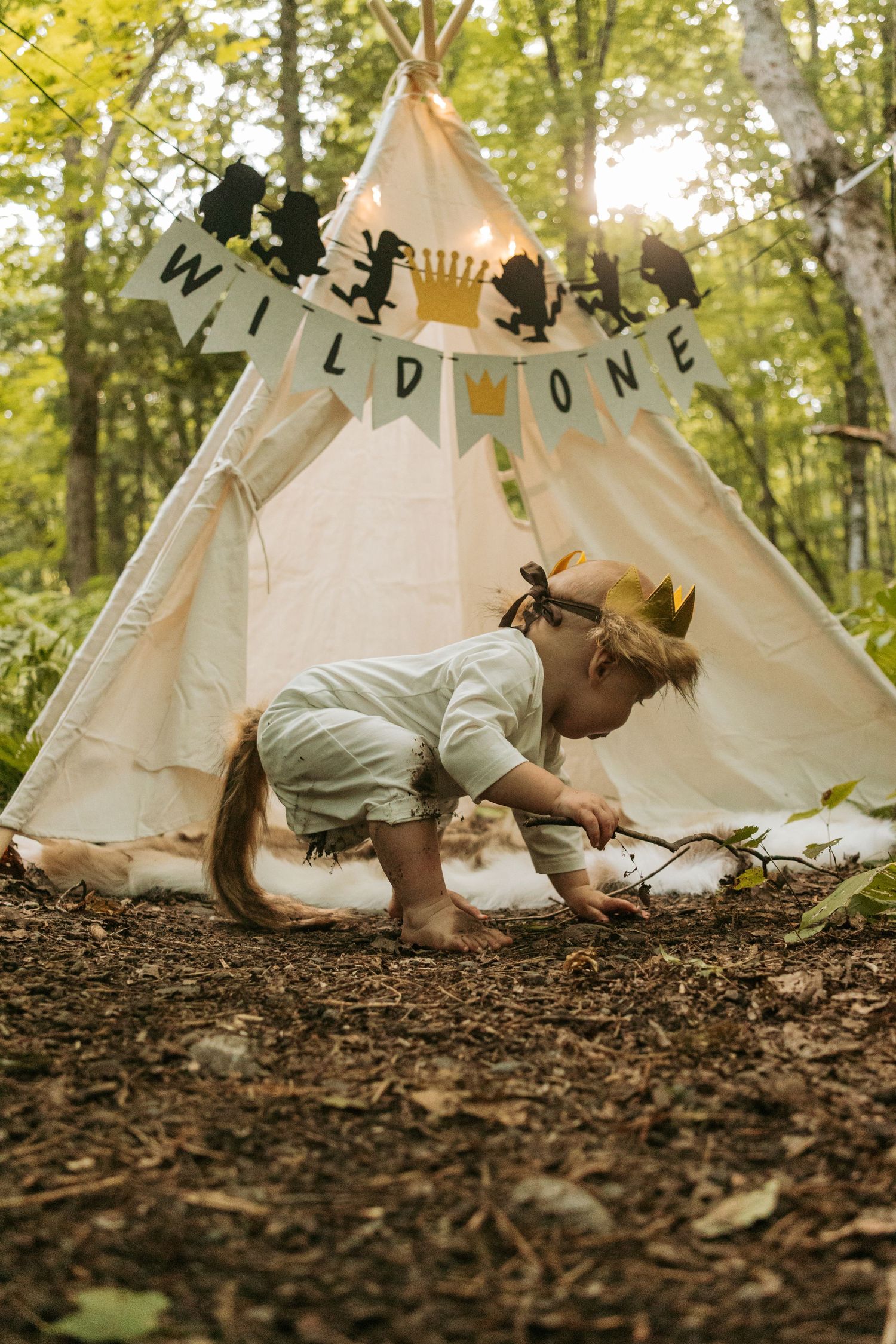 Child exploring ground near decorated teepee tent in wooded area.