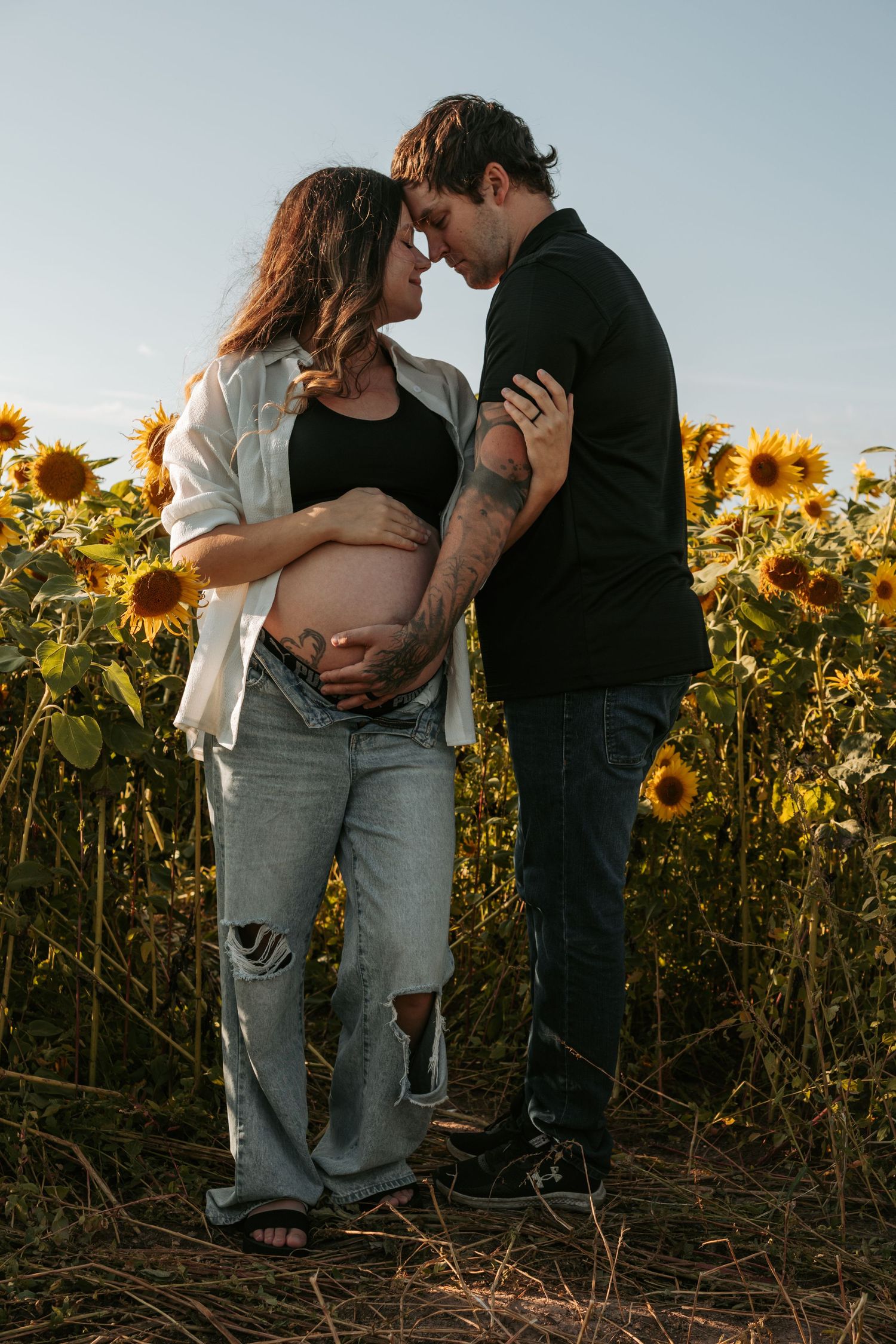 Romantic maternity photoshoot in a field of sunflowers at sunset with couple in casual attire.