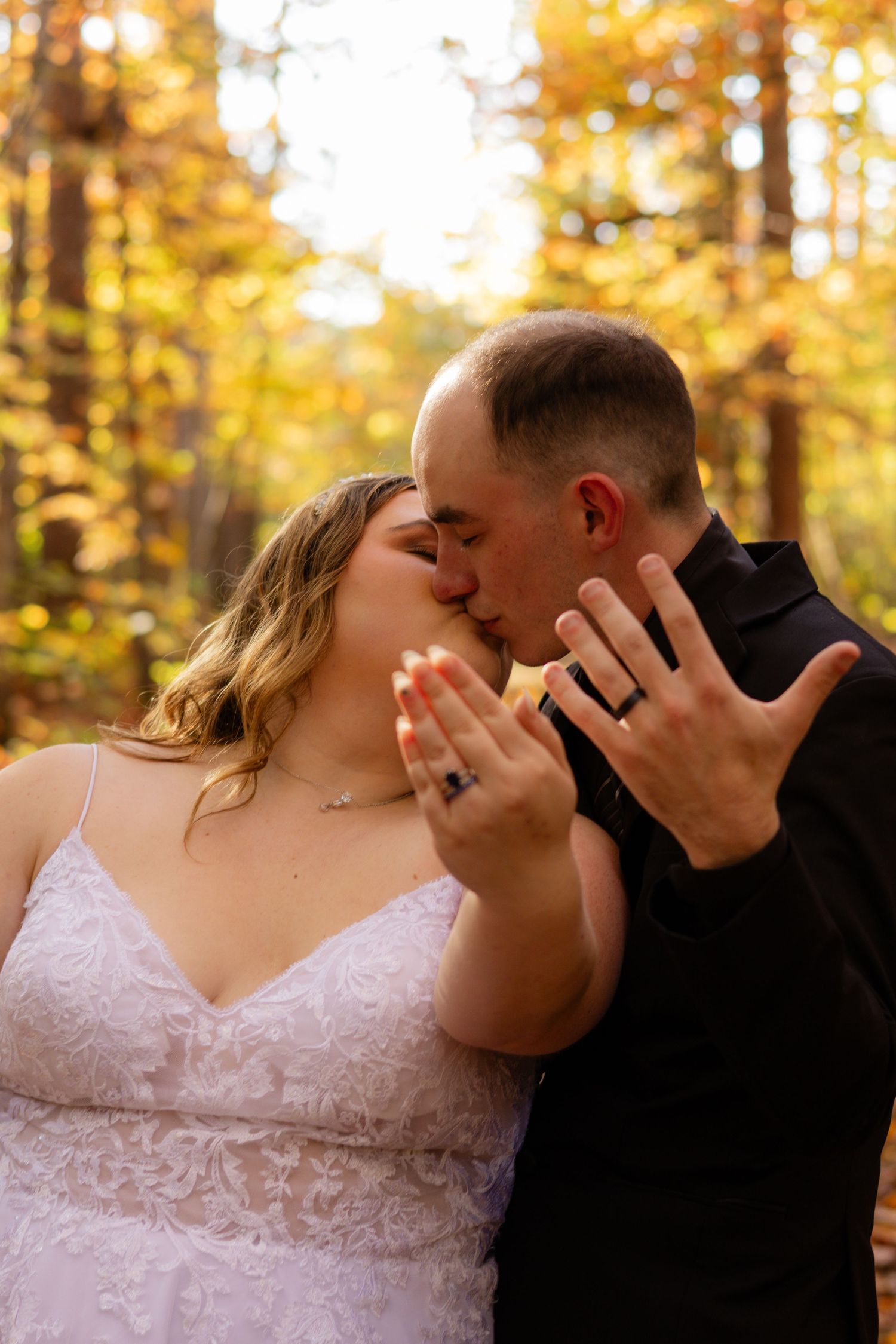 Newlyweds share an intimate kiss while showing off their wedding rings in a sunlit autumn forest.