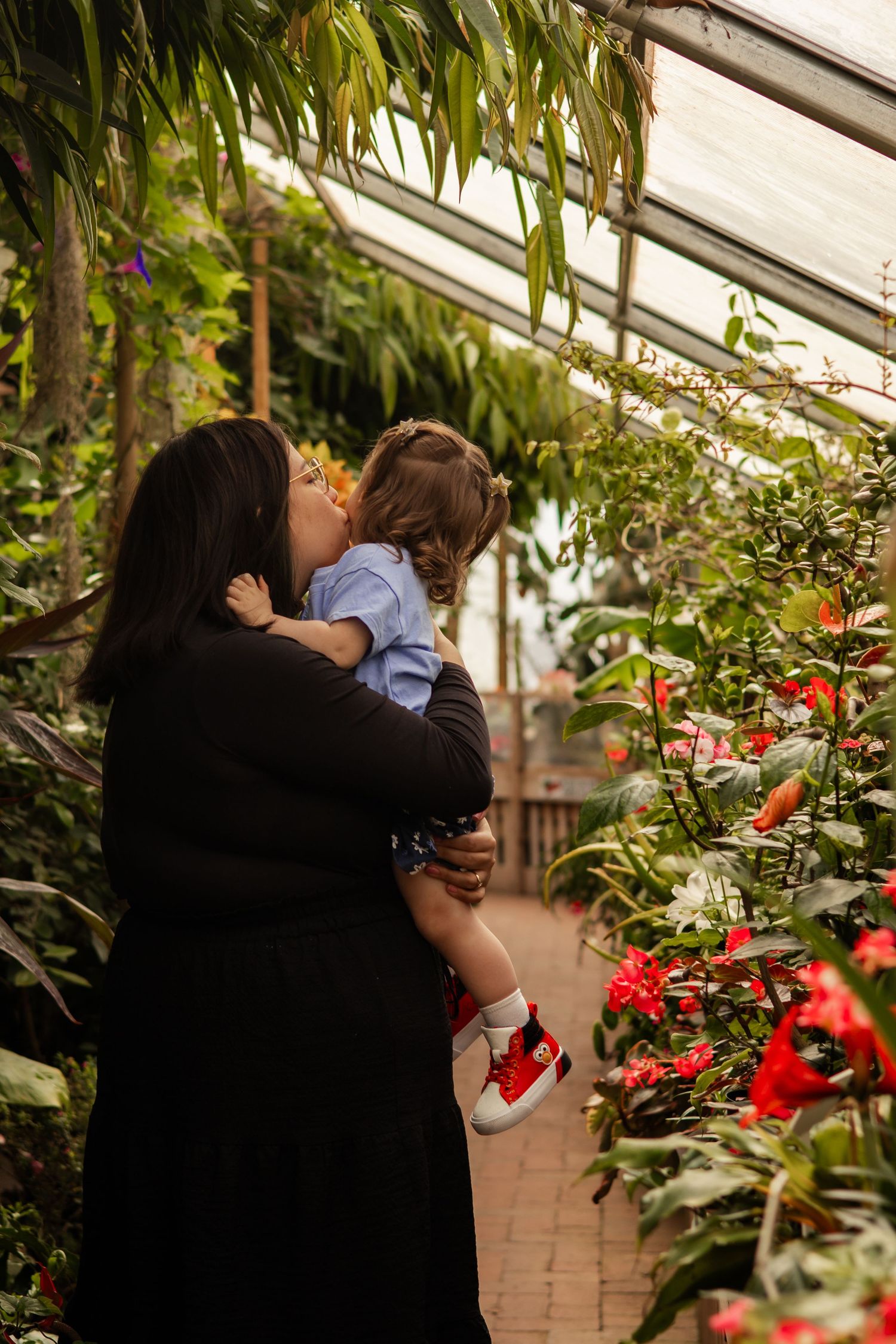 A mother holding and kissing her toddler in a lush greenhouse surrounded by red flowers and green plants.