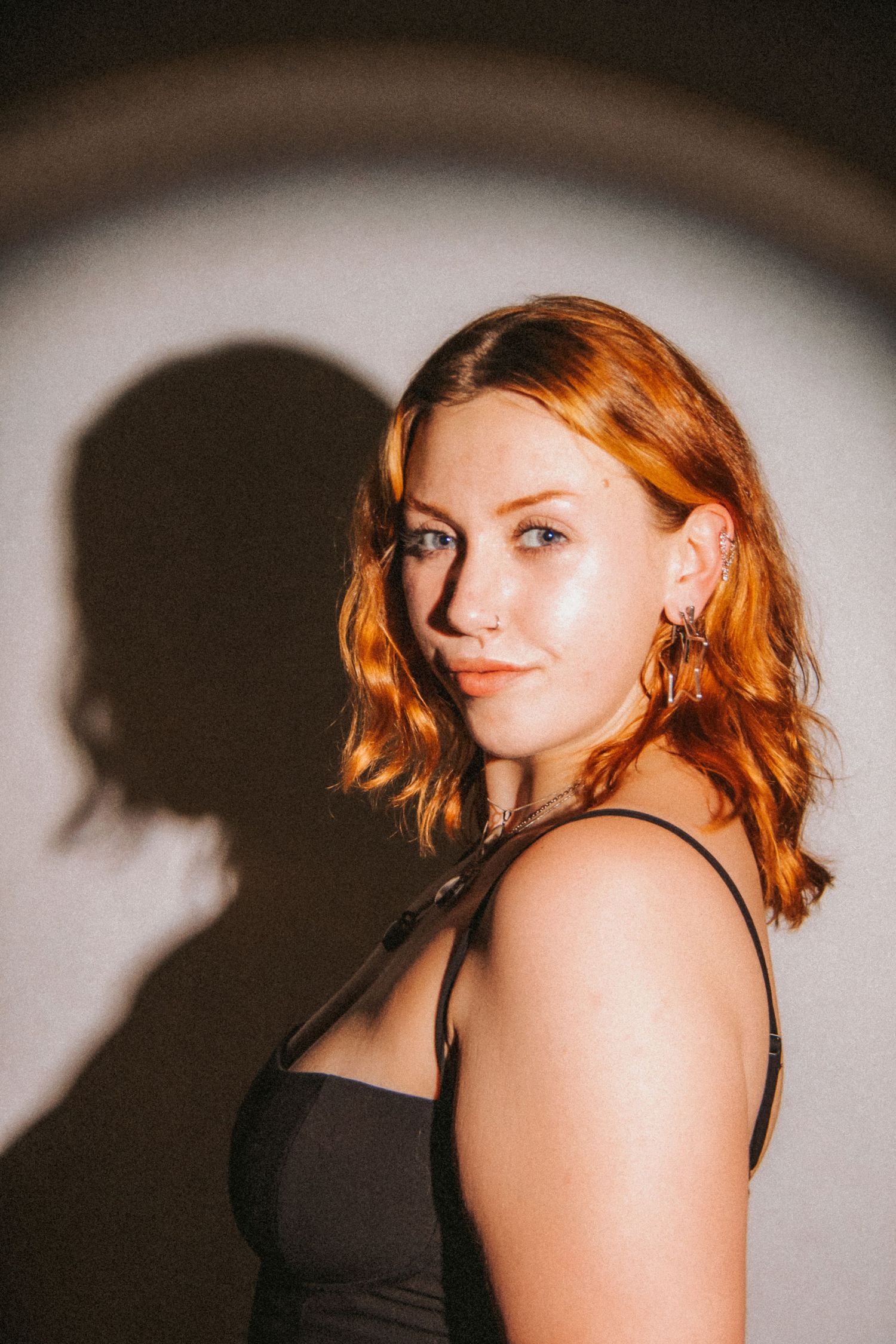 Young woman with red wavy hair wearing a black dress, posing against a white wall with dramatic shadow lighting.