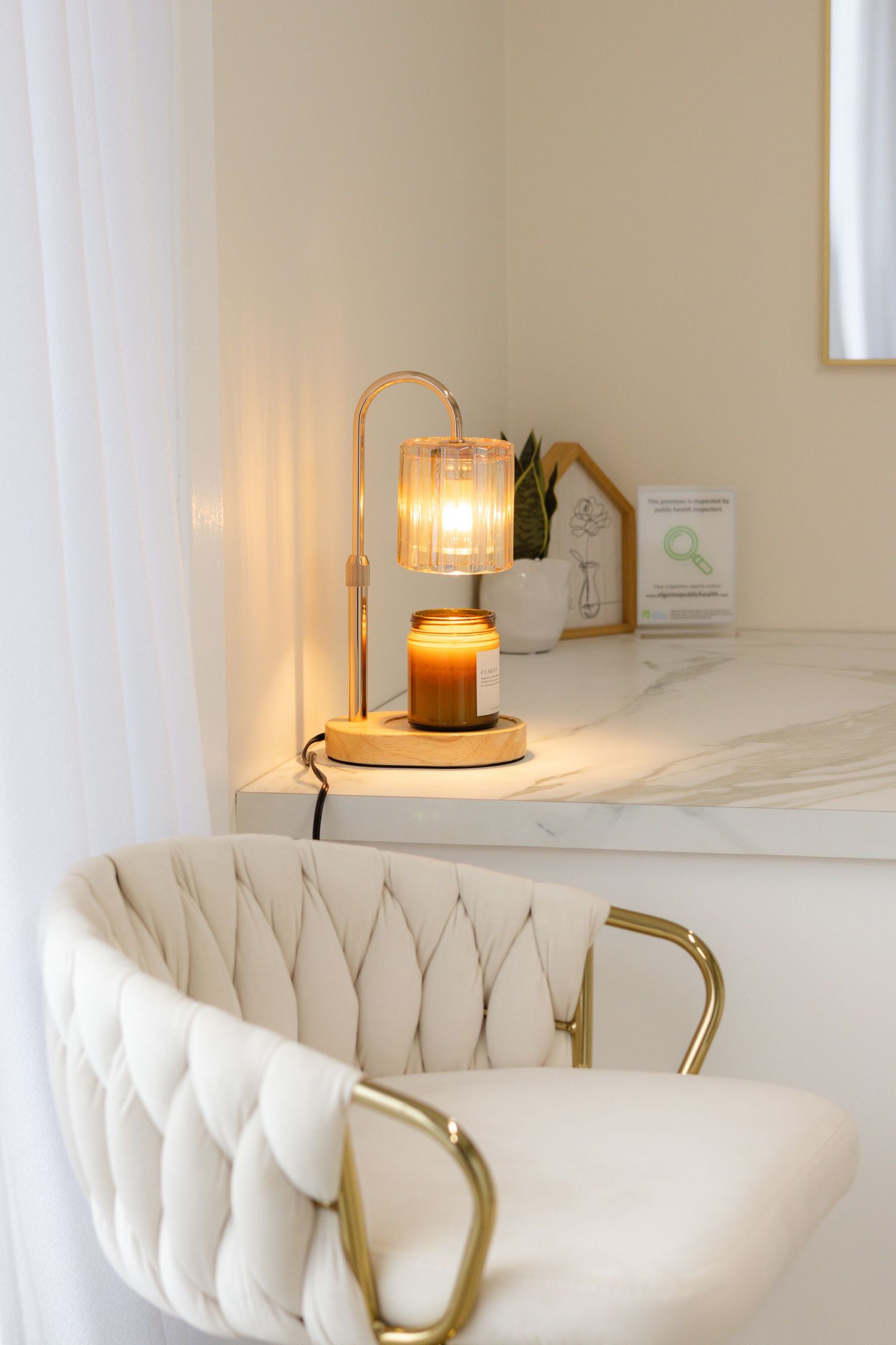 Elegant vanity corner with glowing candle warmer lamp, marble shelf, and tufted white velvet chair with gold arms.