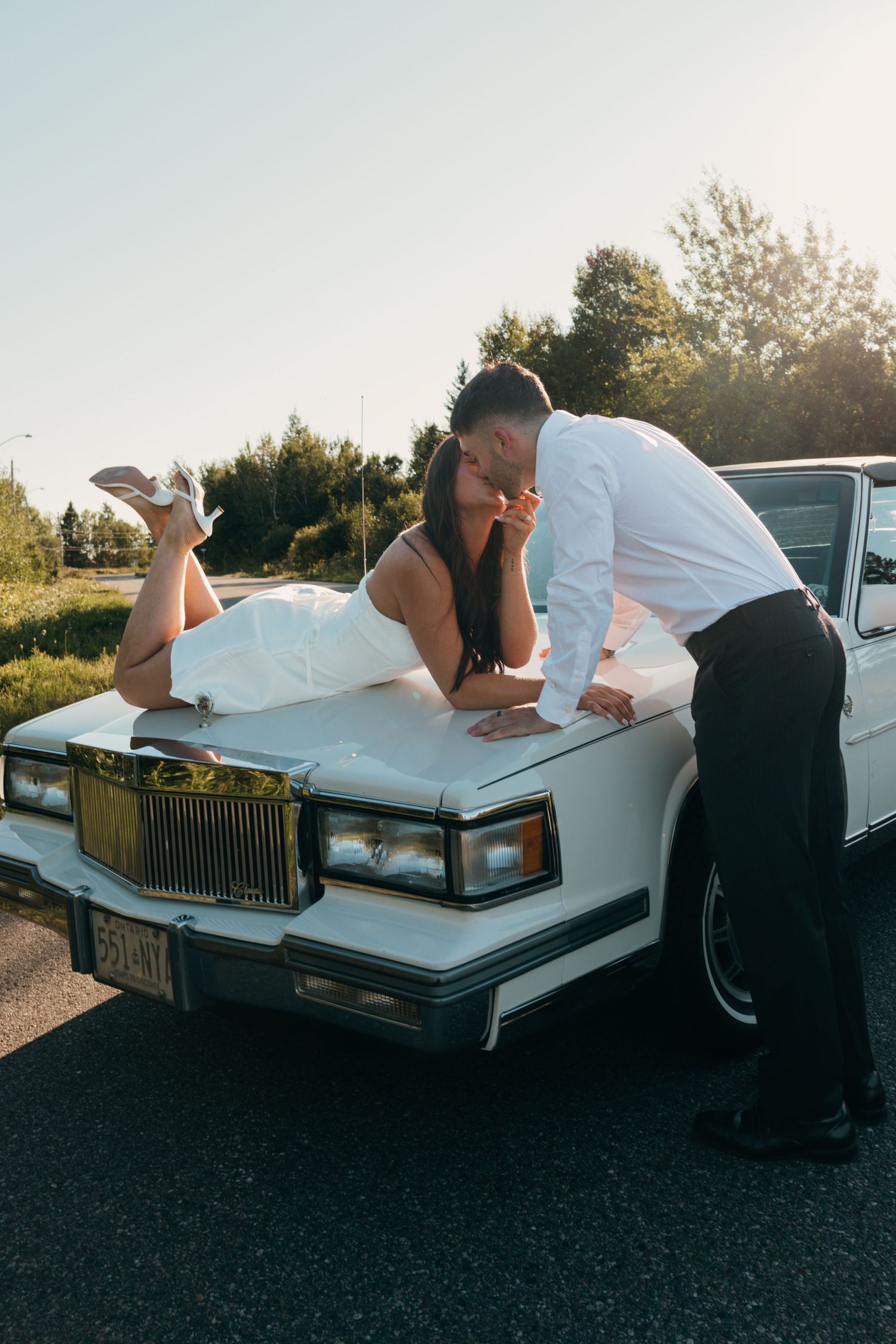 Bride in white dress lying on vintage white car hood kissing groom in white shirt and black pants outdoors.