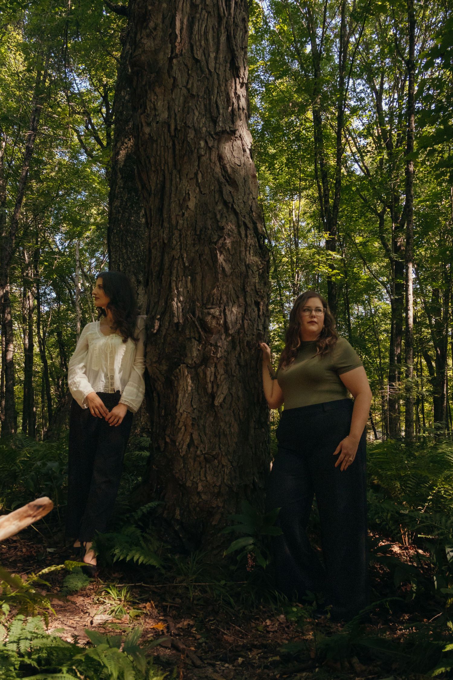Two people stand against a large tree trunk in a lush forest setting.