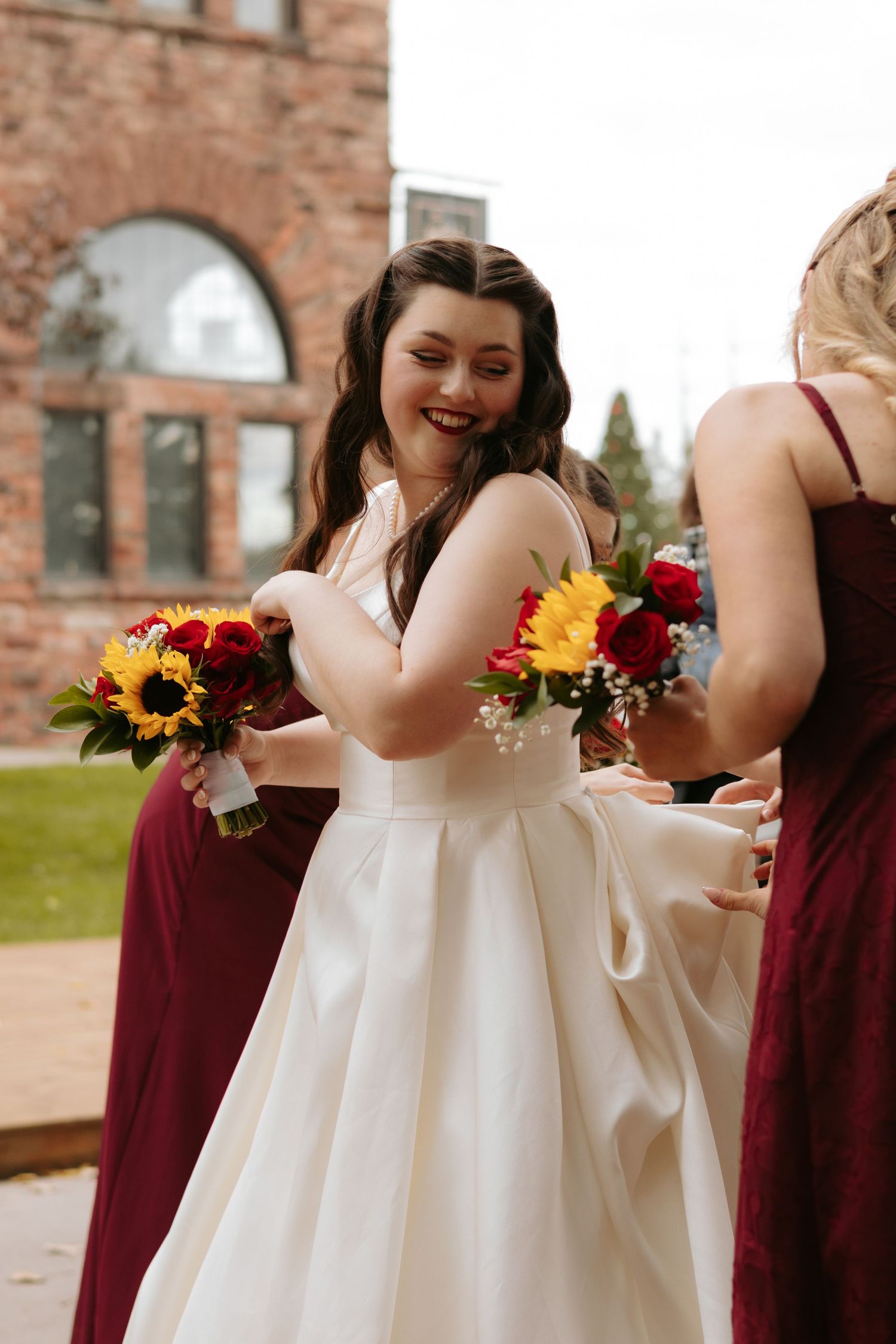Bridesmaids in burgundy dresses hold sunflower bouquets against a brick backdrop.