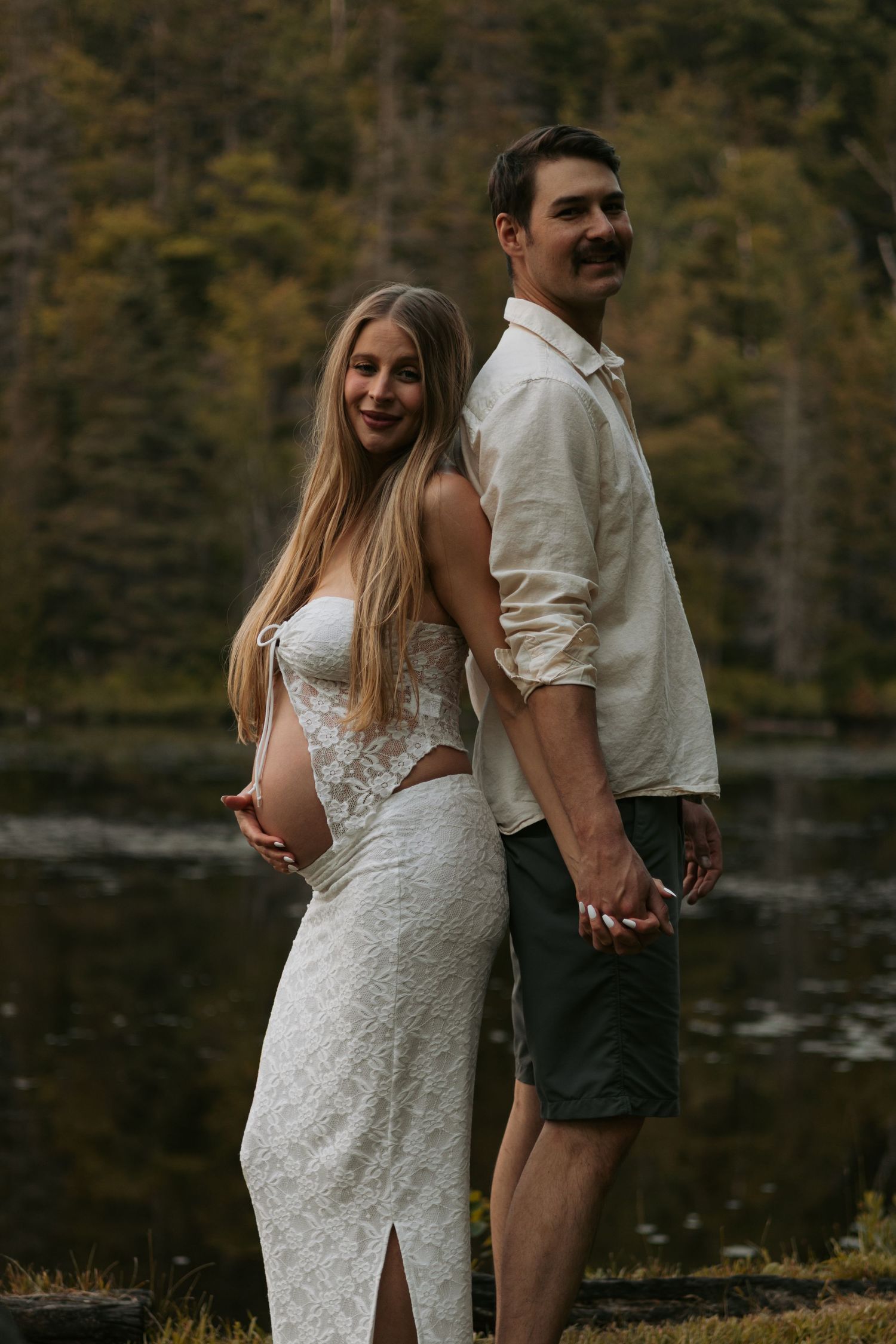 Romantic maternity photoshoot by lake with couple in white lace dress and casual wear.