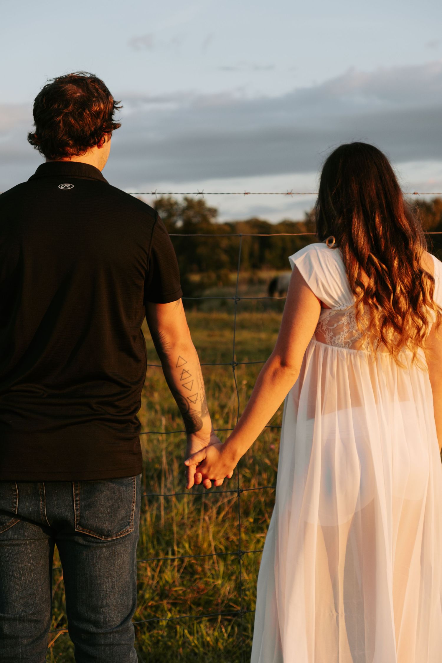 Couple in casual and white dress attire walking through field at sunset holding hands.
