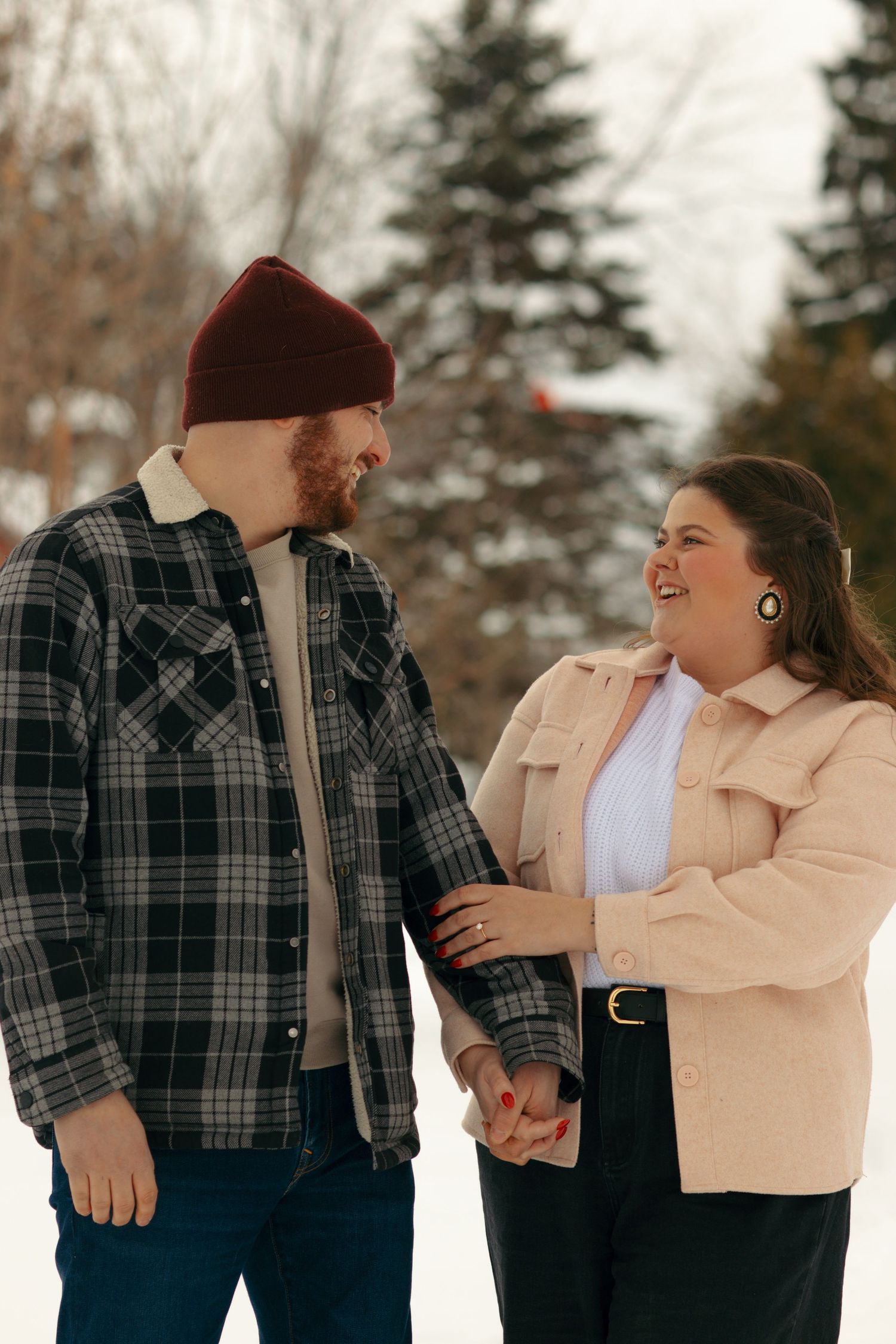 Couple in winter clothing sharing a joyful moment in the snow with evergreen trees in background.