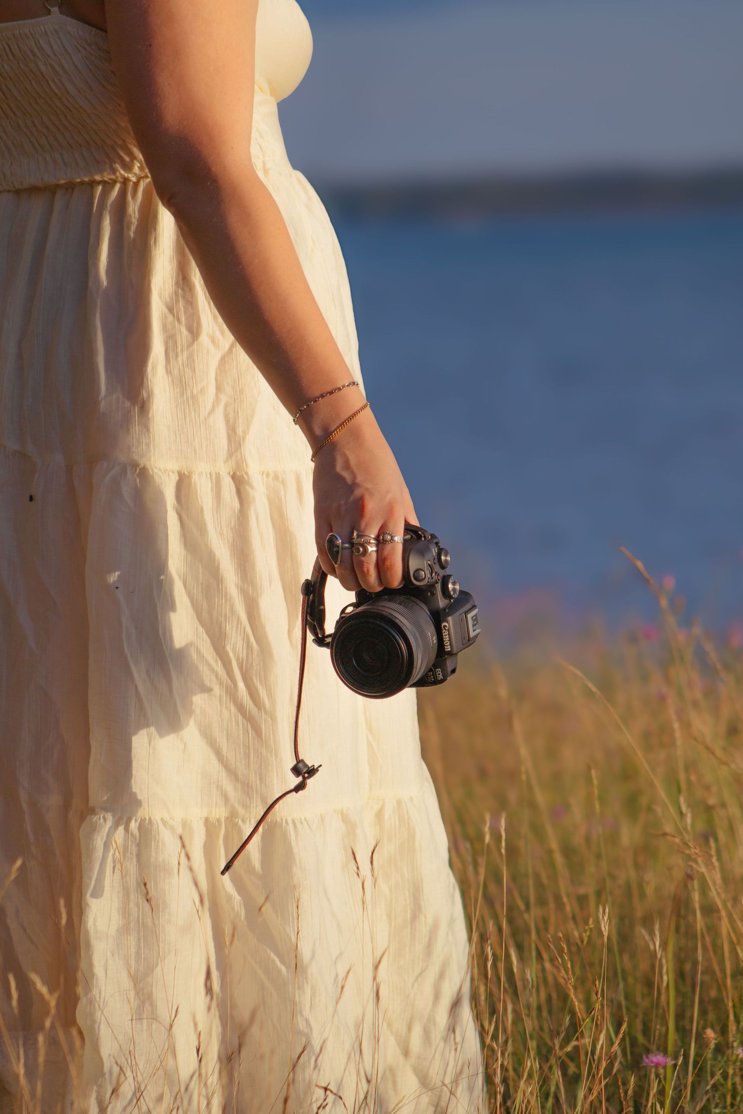 Woman in white dress holding a camera by a lakeside field at golden hour.