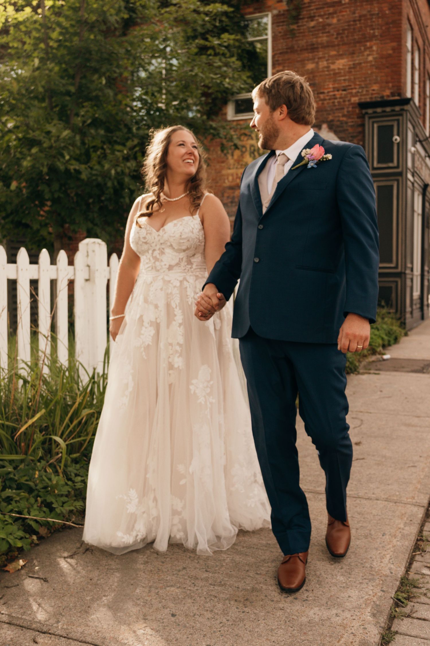 A bride in a flowing lace gown walks with her groom along a brick building pathway.