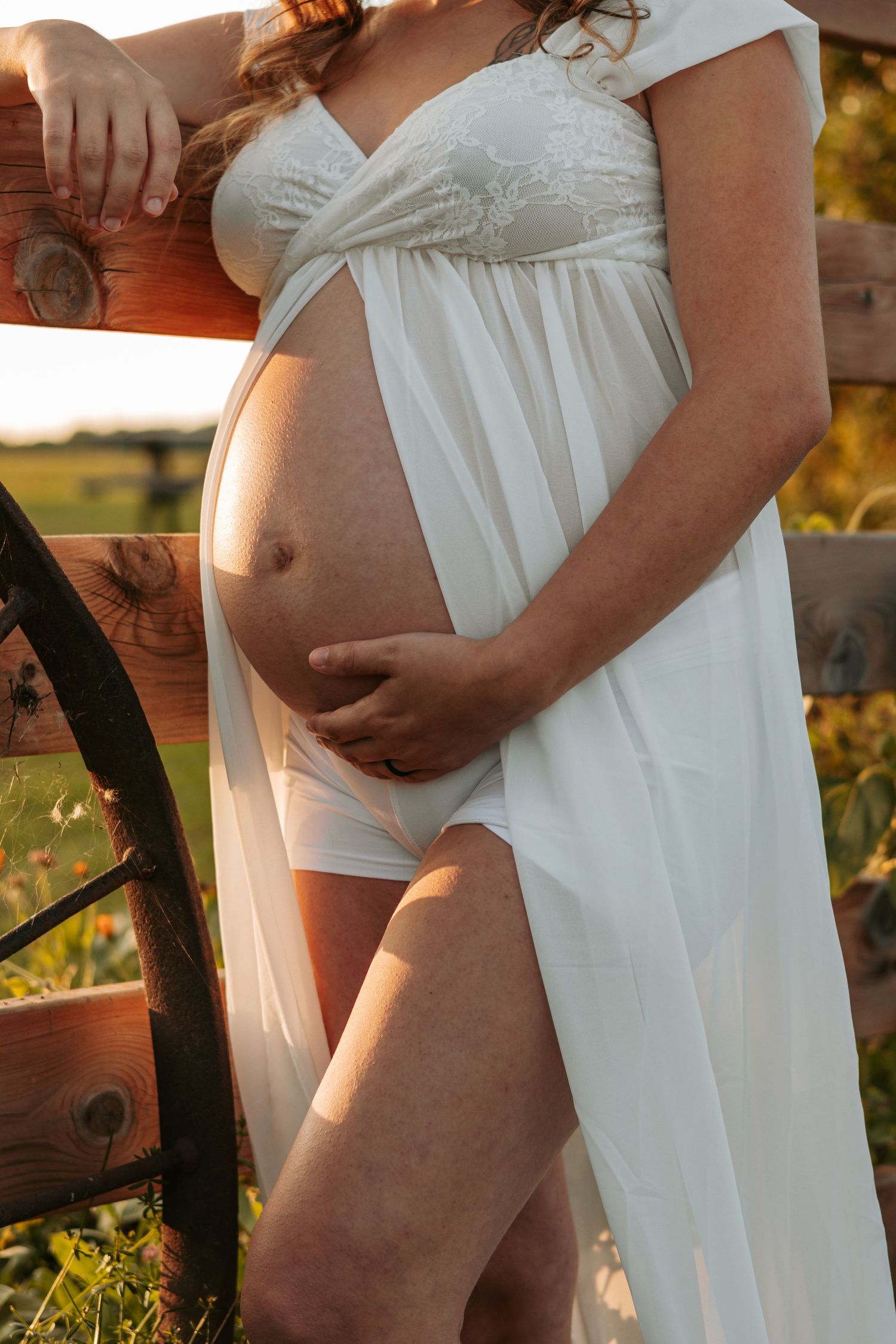 Artistic pregnancy photos in white dress against rustic backdrop at sunset.