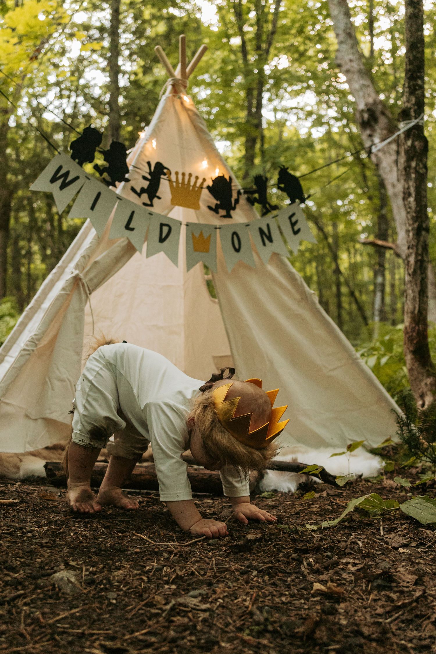 Toddler crawling on forest floor near decorated teepee tent with banner.