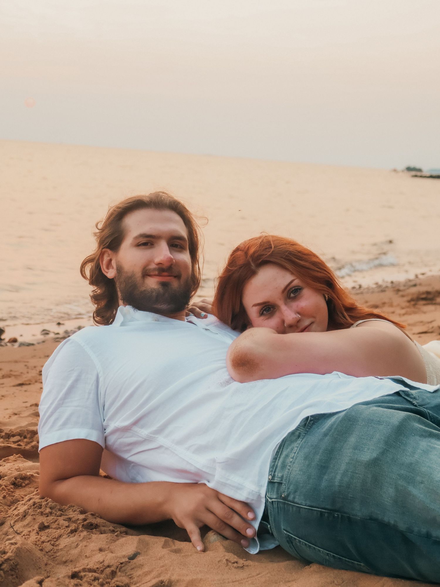 Couple relaxing on sandy beach at sunset, woman with red hair resting on man's chest, both smiling.