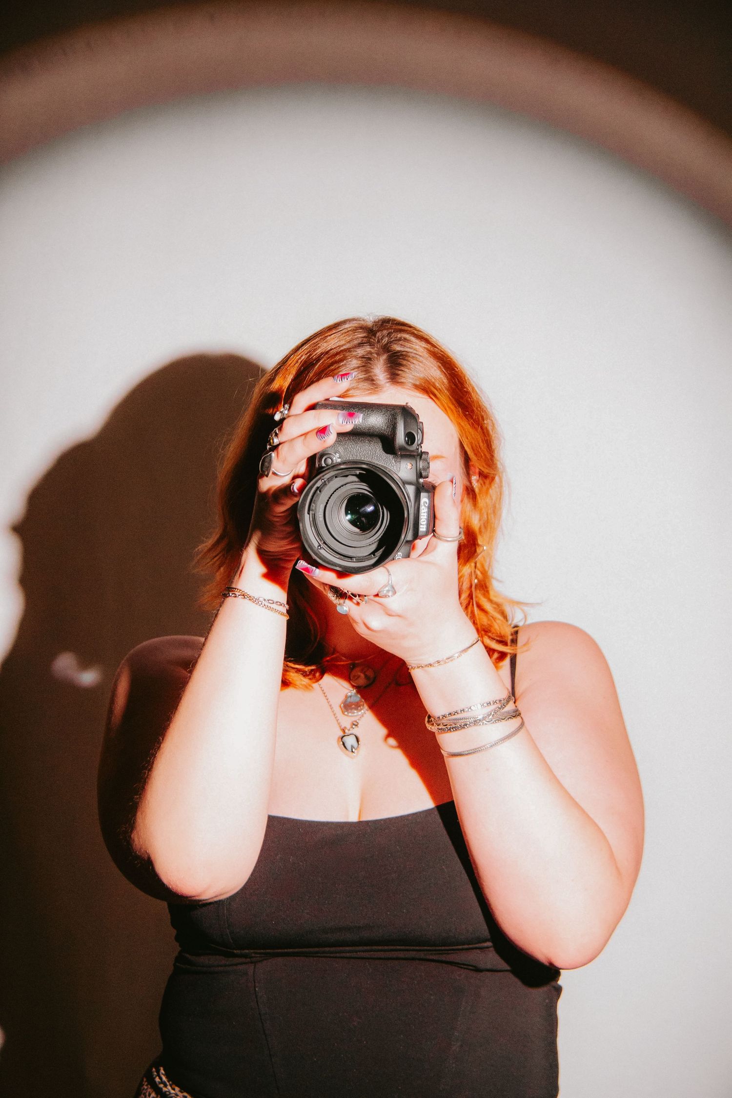 A redhead woman in a black top holds a DSLR camera up to her face, standing against a white wall with circular light.