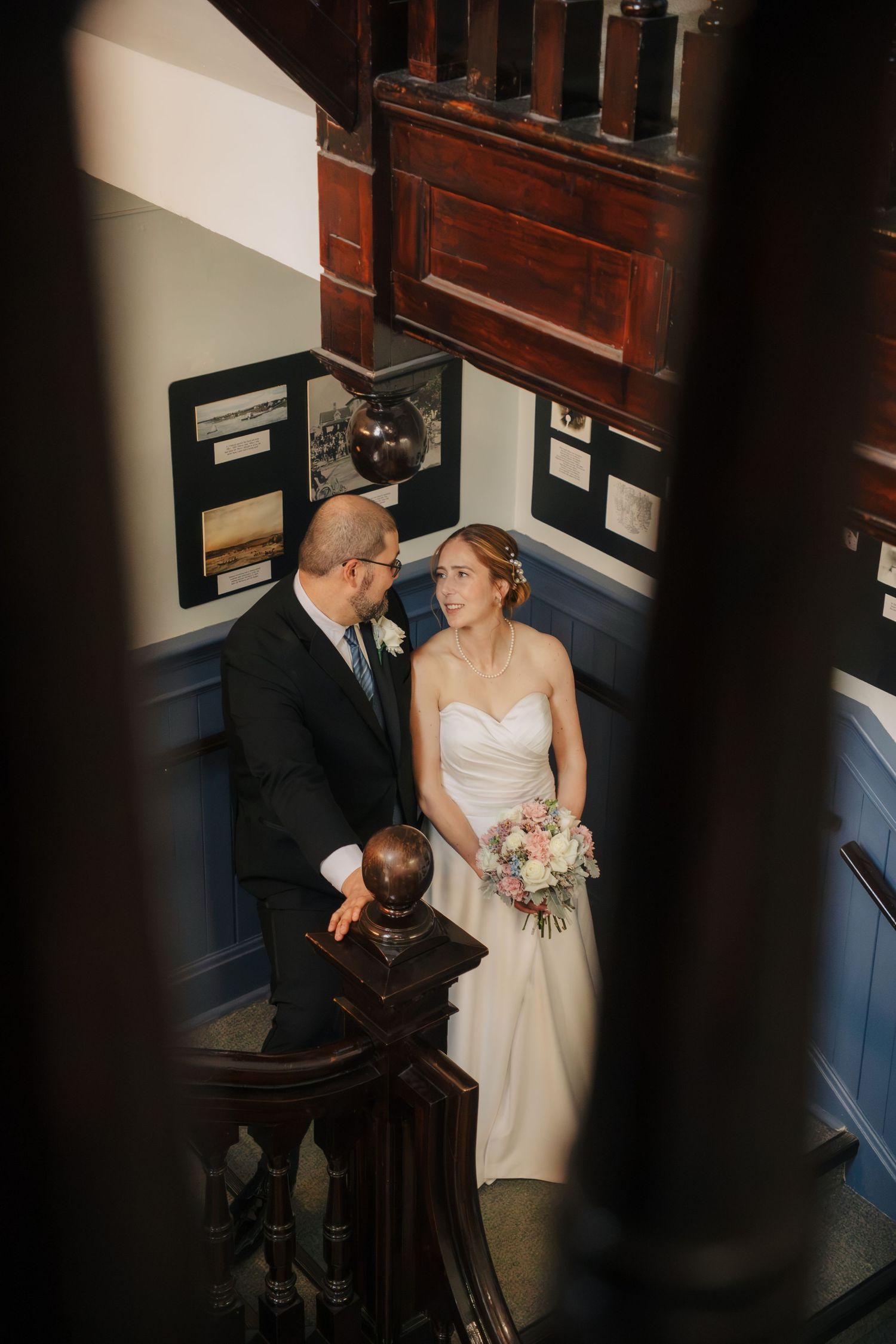 Bride and groom share a tender moment on a staircase, bride holding a bouquet in a white gown at their wedding venue.
