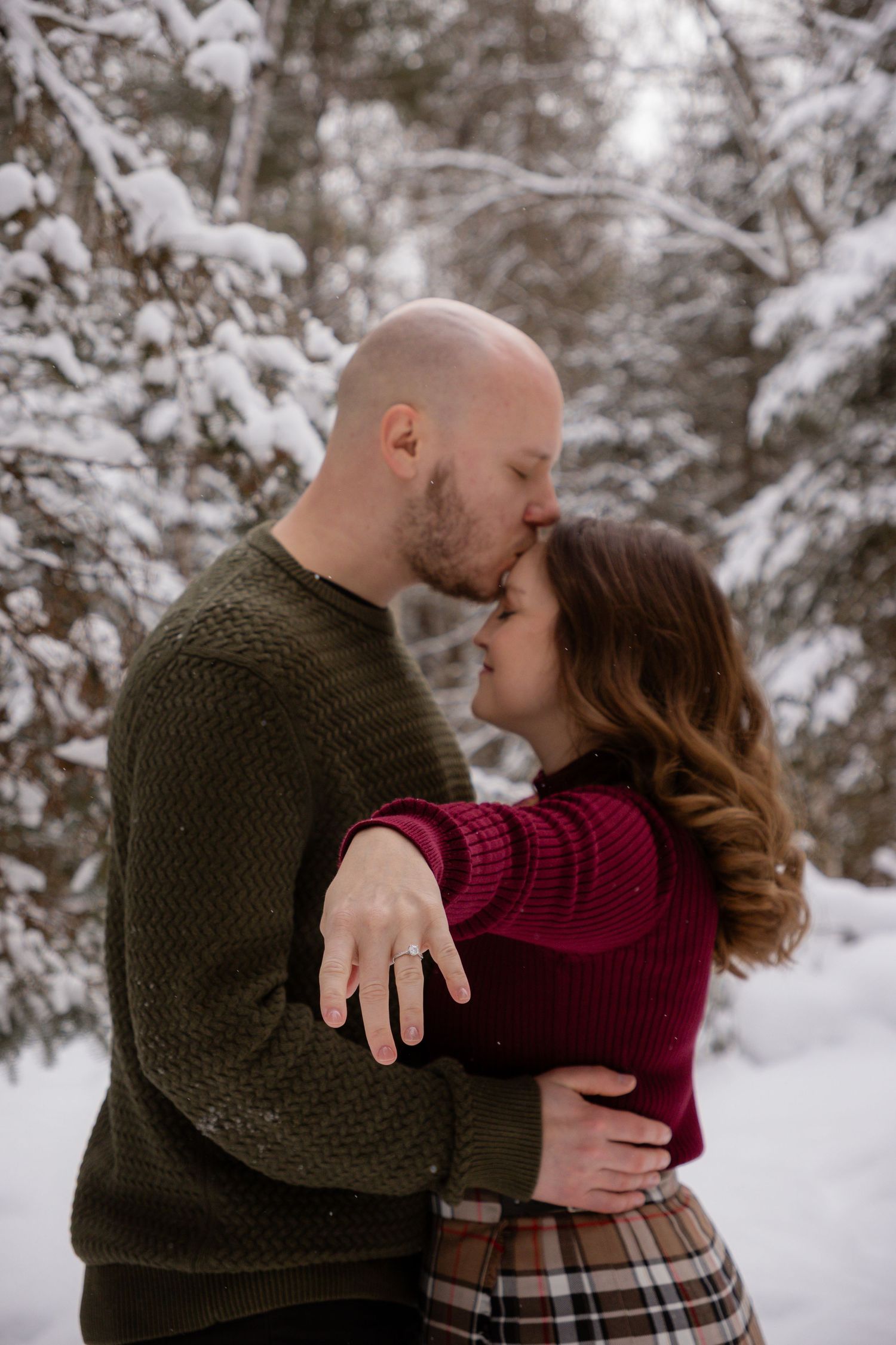 A romantic winter engagement photo showing a couple embracing in the snow with a sparkling ring on display.