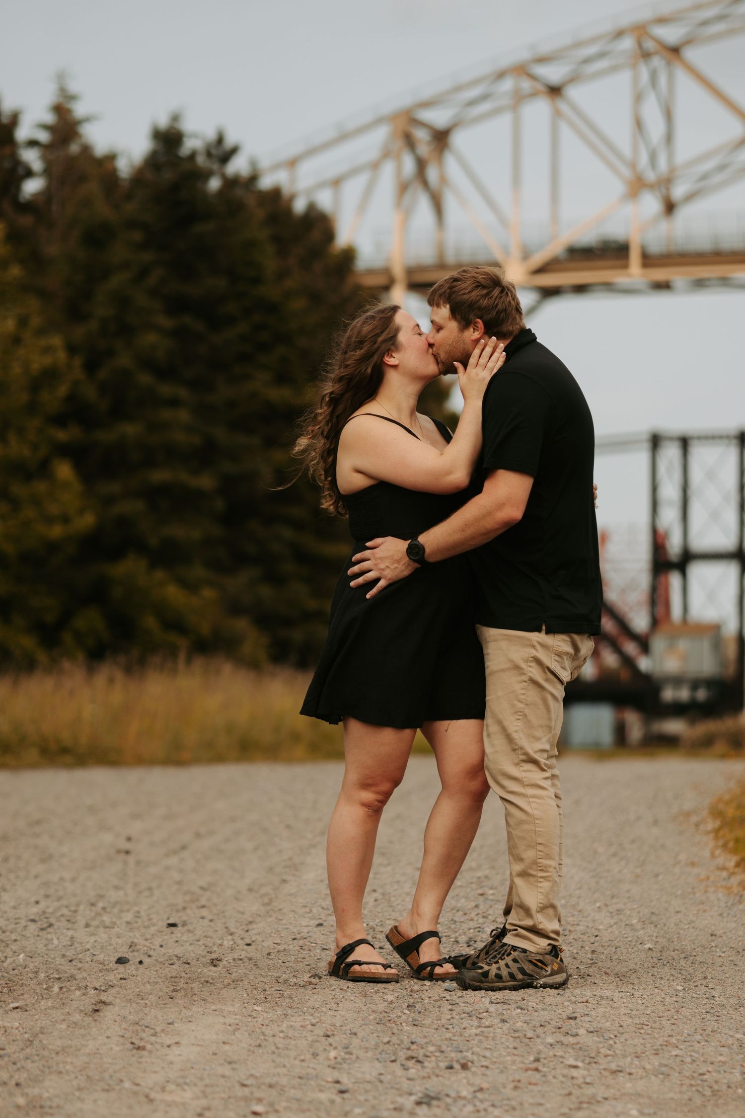 Romantic couple sharing intimate moments on a sandy path with industrial bridge in background.