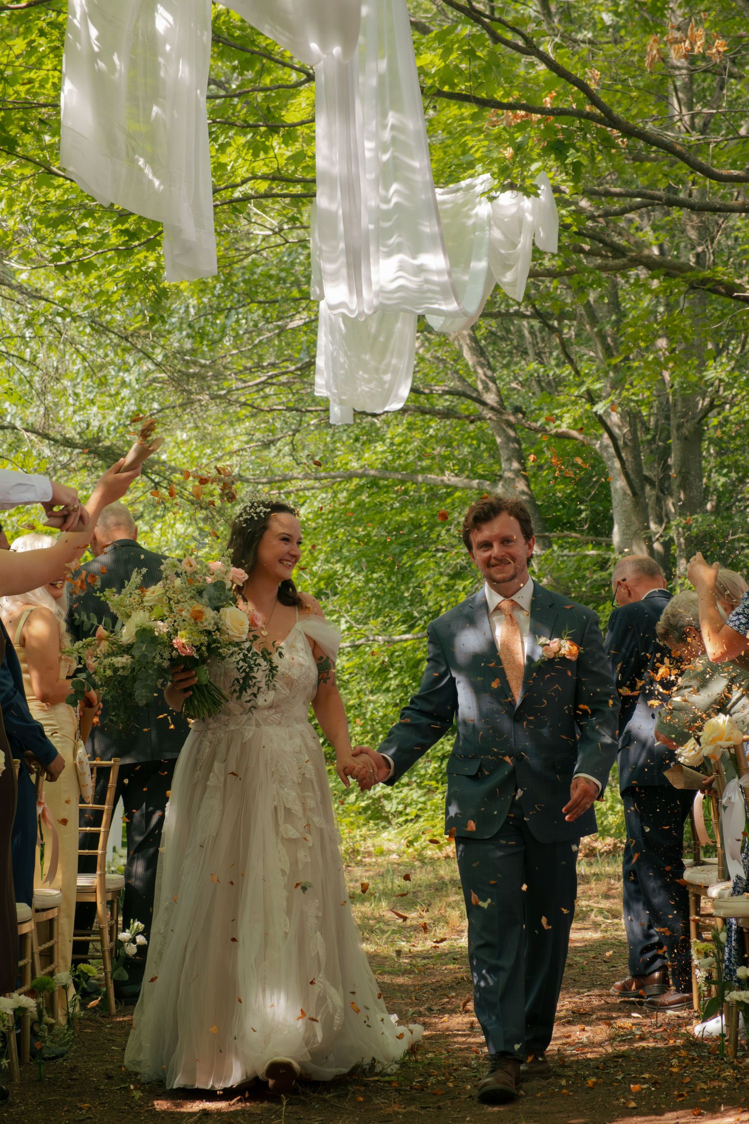 Newlywed couple walks hand-in-hand through confetti shower in woodland wedding ceremony aisle.