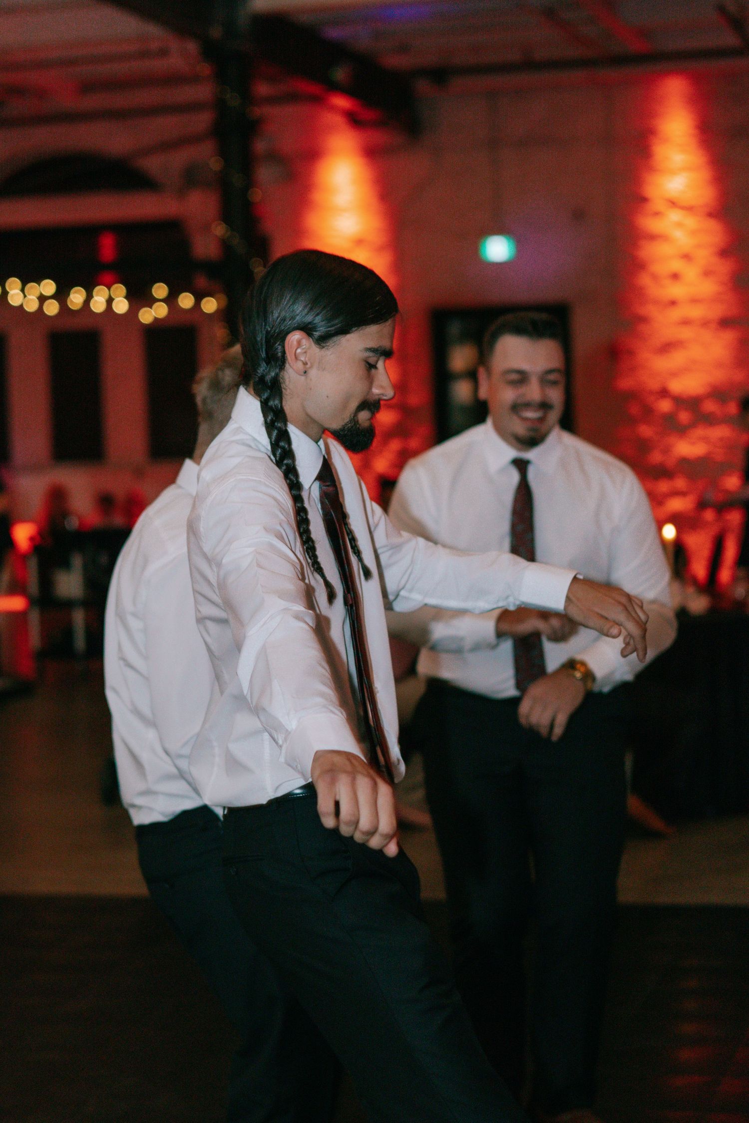 Guests dancing energetically at a wedding reception with warm red lighting and string lights overhead.
