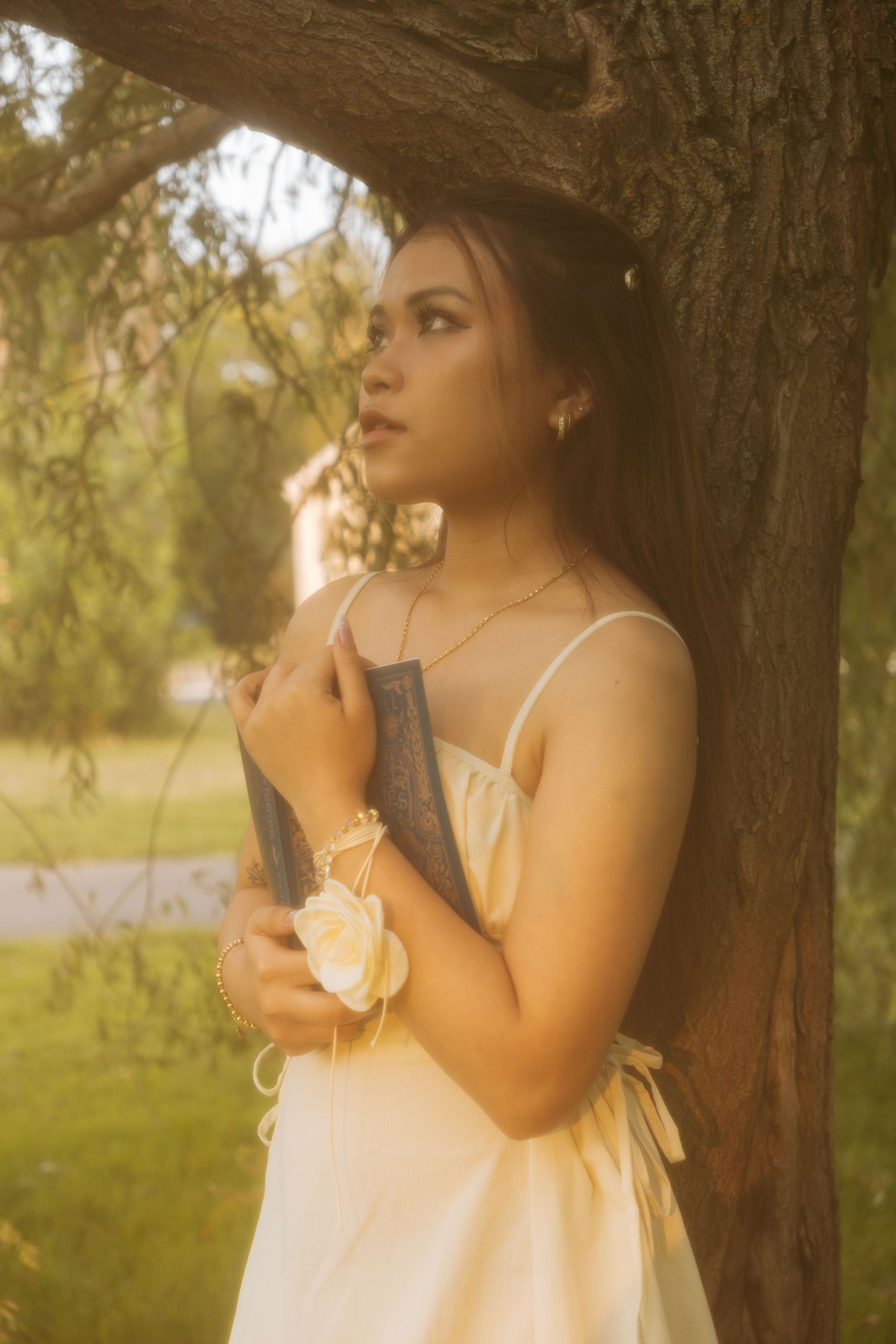 Woman in white dress poses thoughtfully by a tree in golden light.