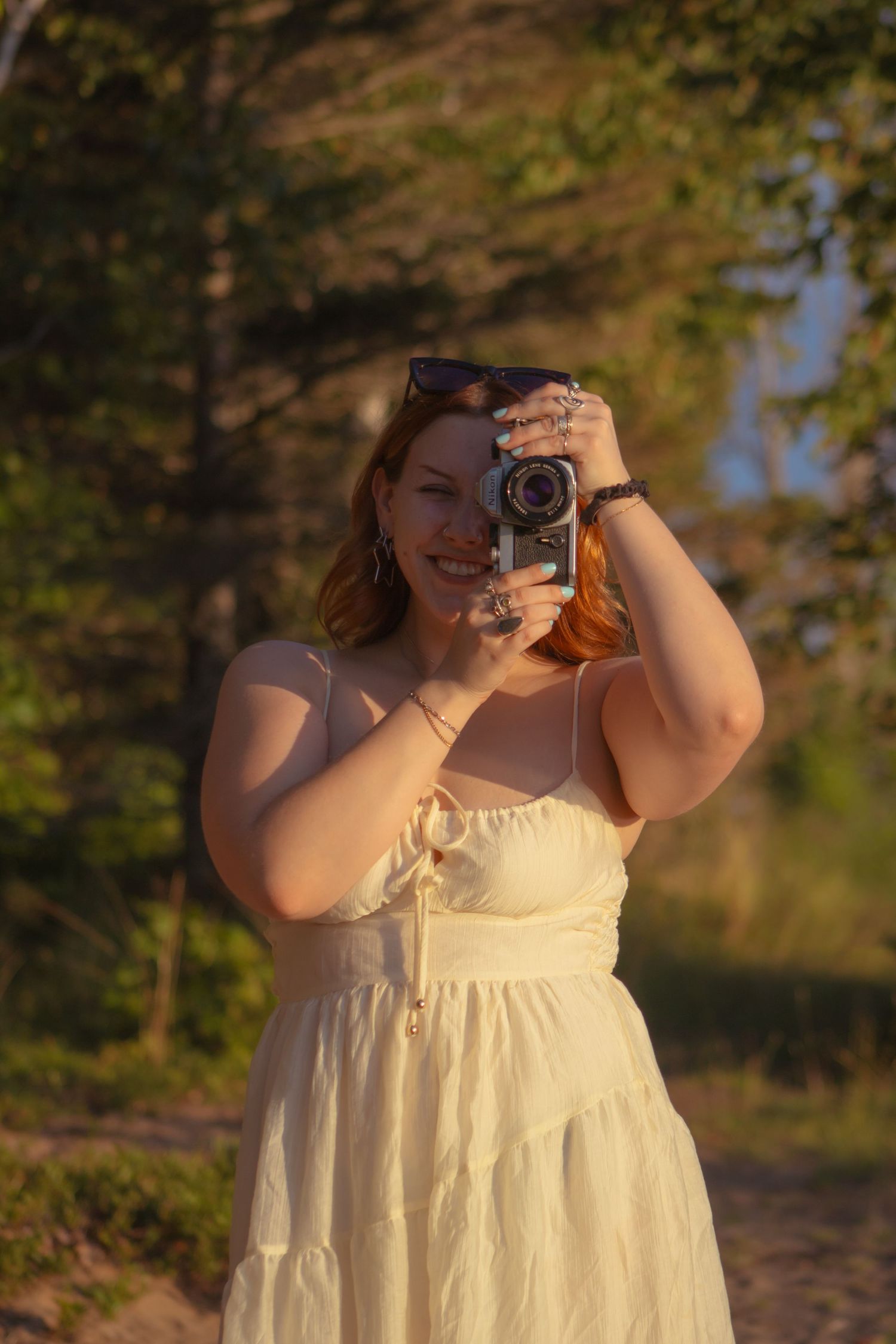 A young redhead woman in a white sundress holds a vintage camera outdoors during golden hour.