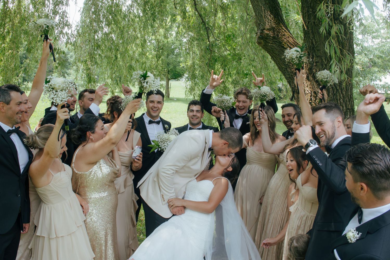 Wedding party celebrating outdoors under willow tree, bride and groom kissing surrounded by cheering bridesmaids and groomsmen.