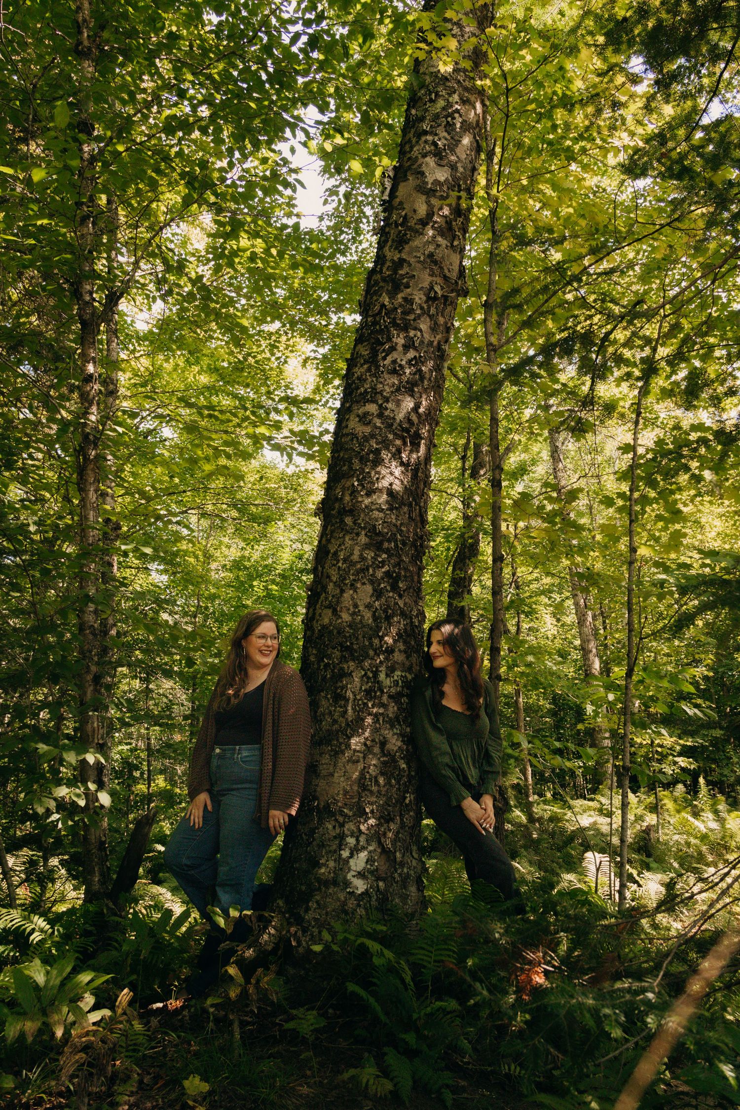 Two people pose on opposite sides of a tall tree trunk in a green forest.