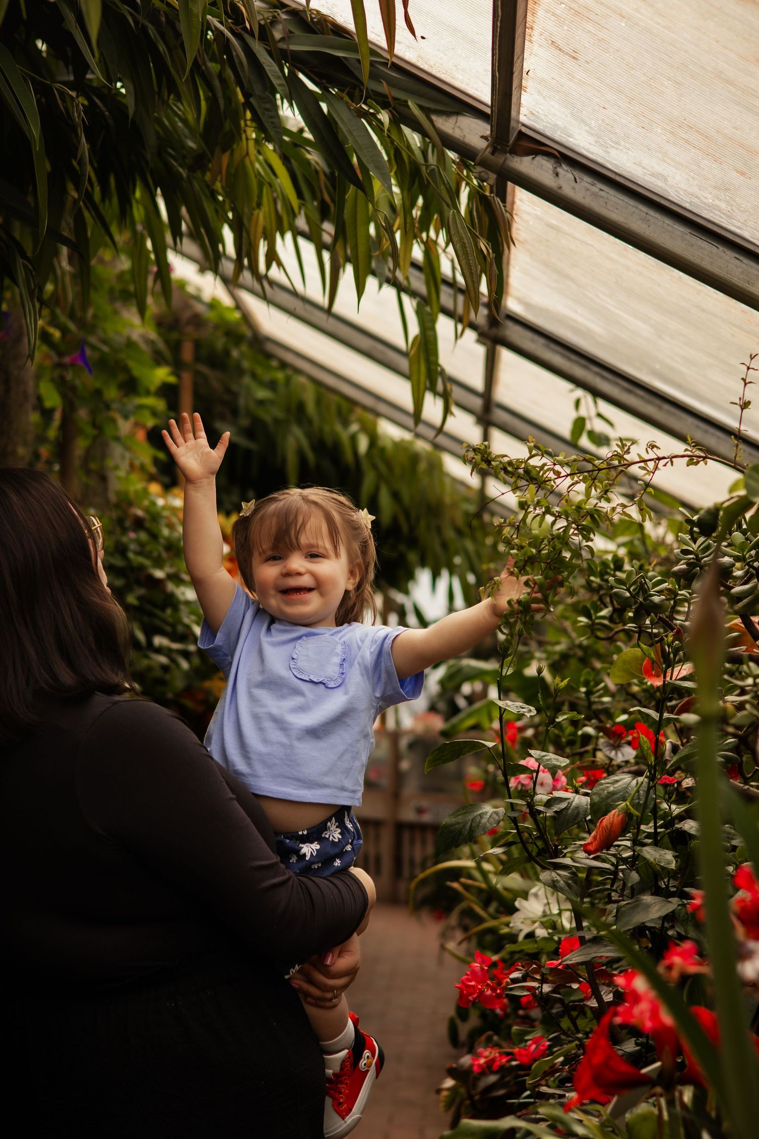Excited toddler girl with arms raised being held by mother among vibrant greenhouse flowers.