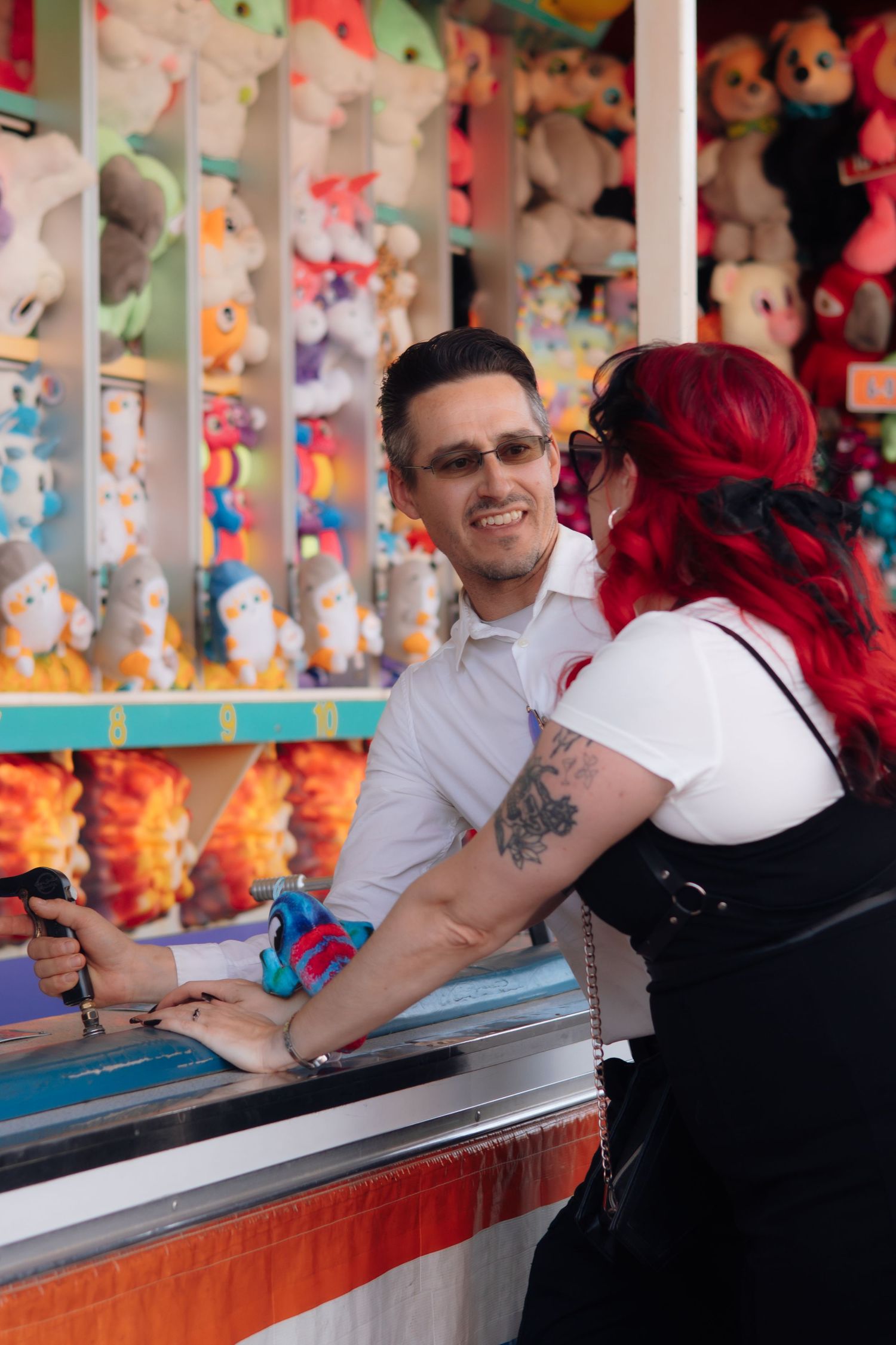A couple smiles at a colorful carnival game booth filled with stuffed animal prizes.