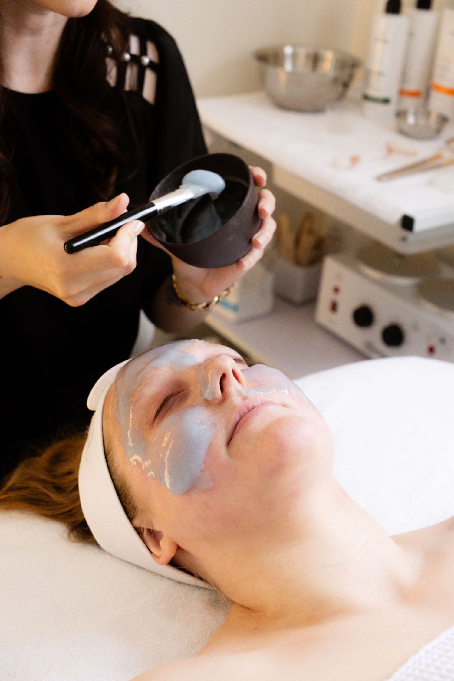 Esthetician applying gray clay facial mask with brush on relaxed woman lying in spa treatment room.