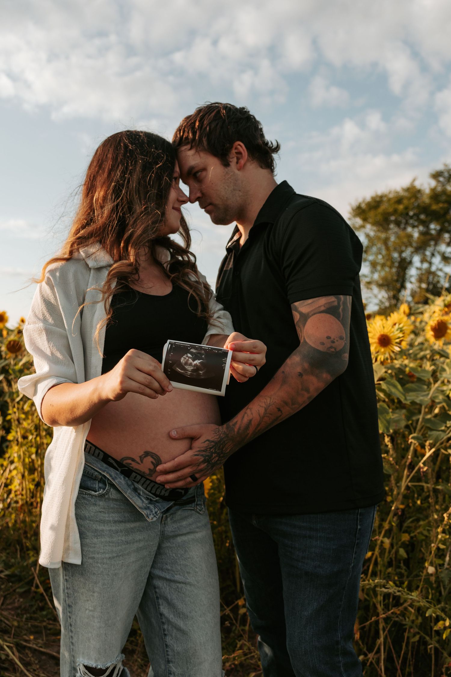 Expectant couple holds ultrasound photo while embracing in golden sunflower field.