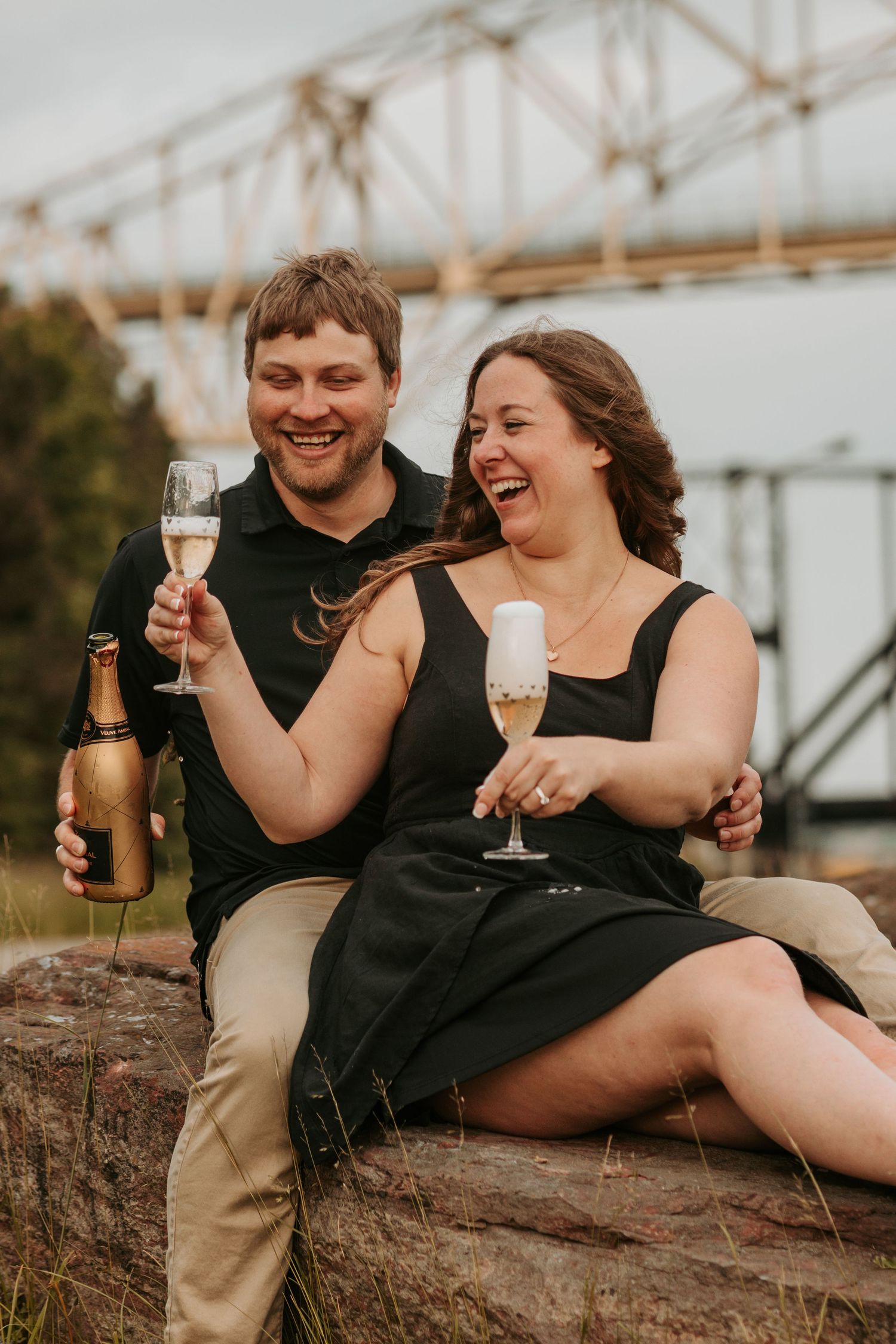 Couple enjoying champagne and laughter while sitting on rocks near industrial bridge.