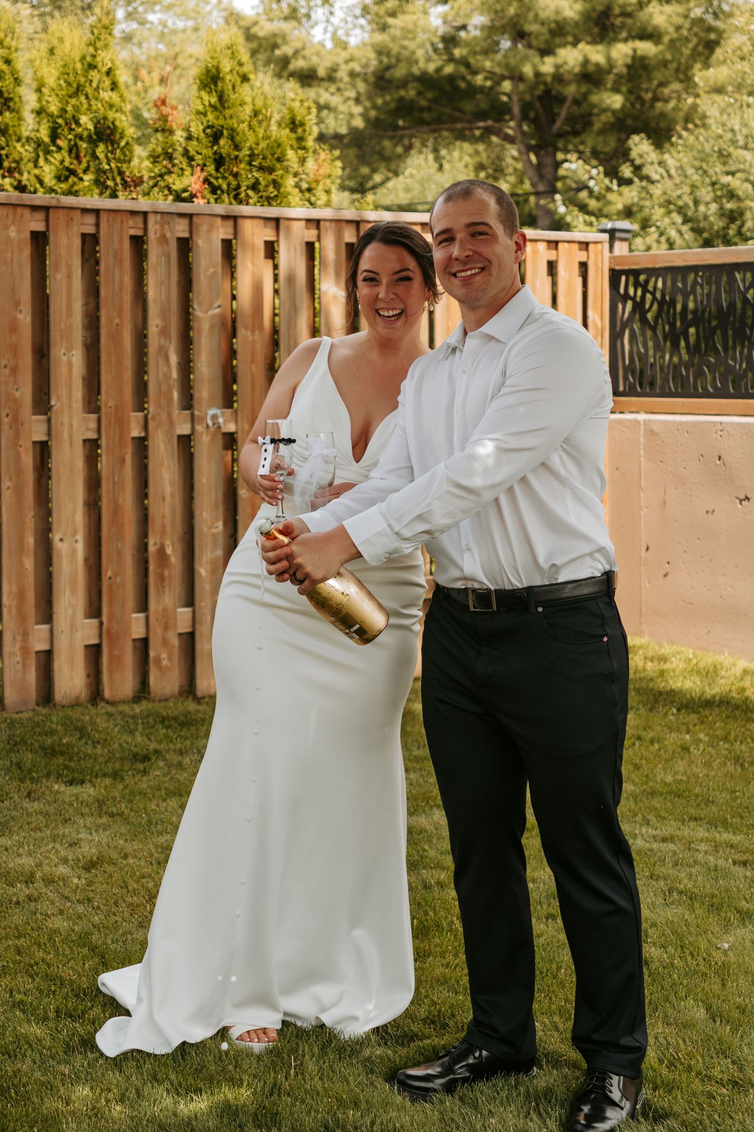 A couple in wedding attire shares drinks and laughter in an outdoor garden setting.