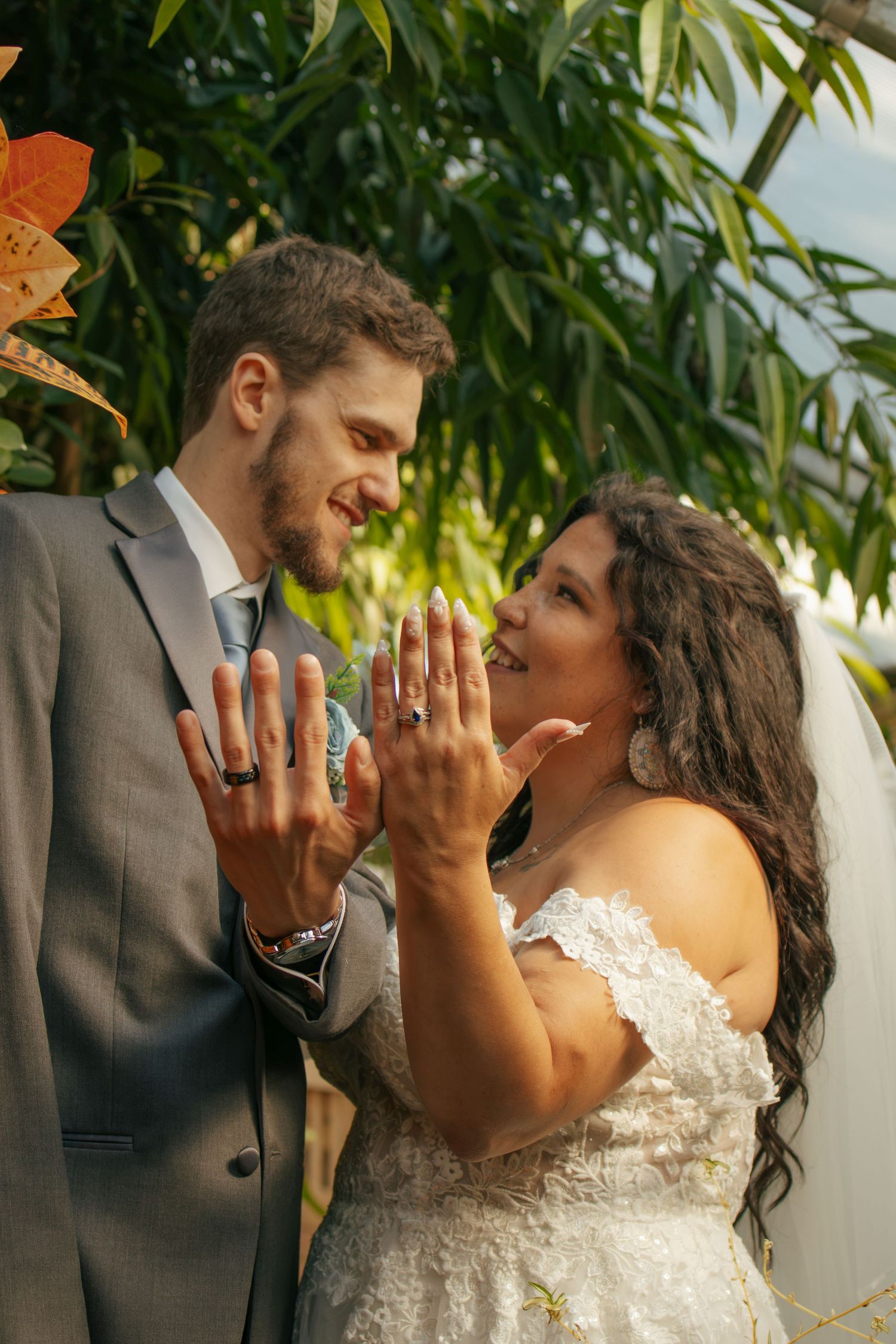 Newlywed couple smiling and showing off wedding rings outdoors, bride in lace gown and groom in gray suit.