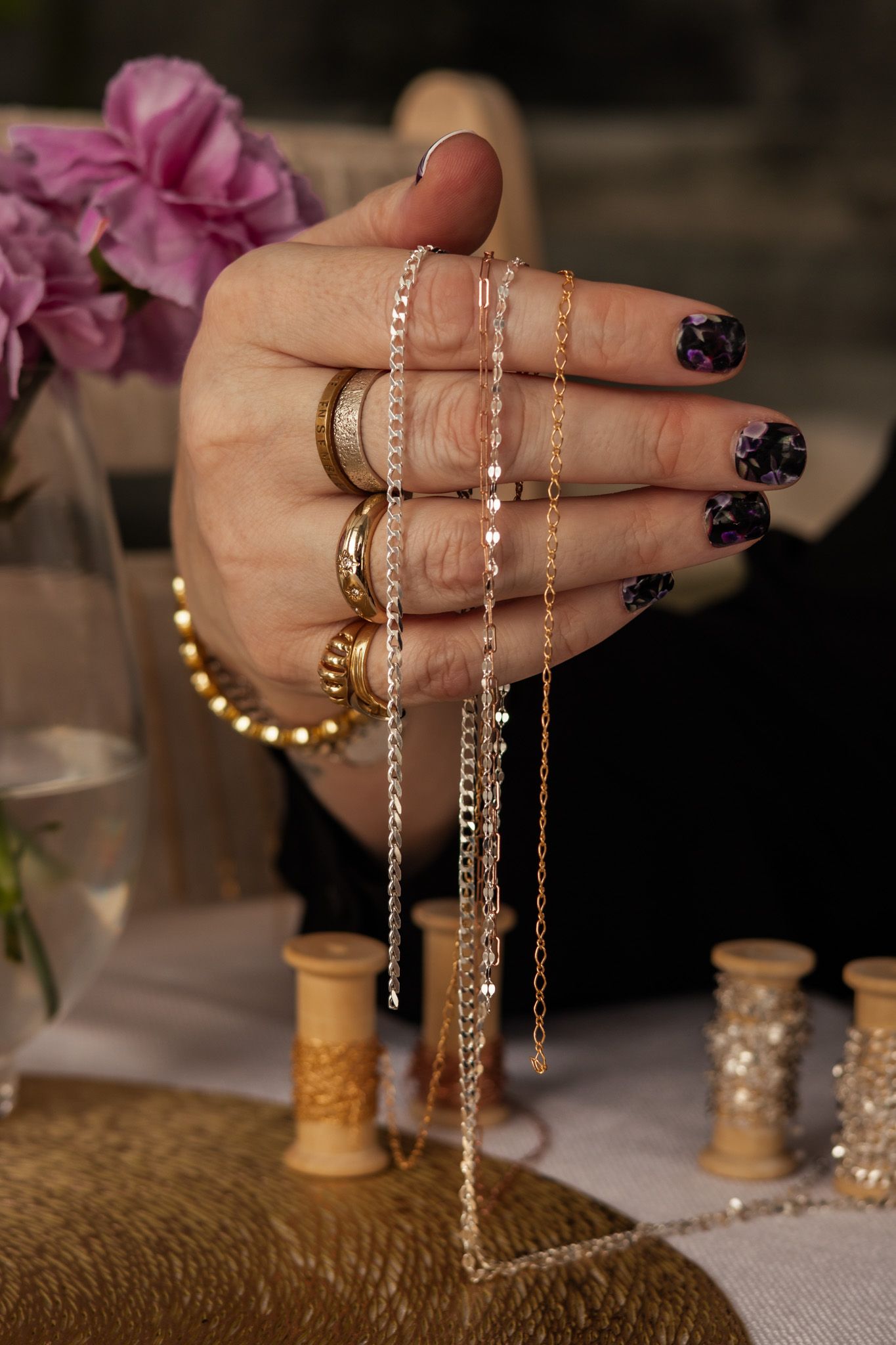 Manicured hands displaying various gold and silver chains against a textured table setting.