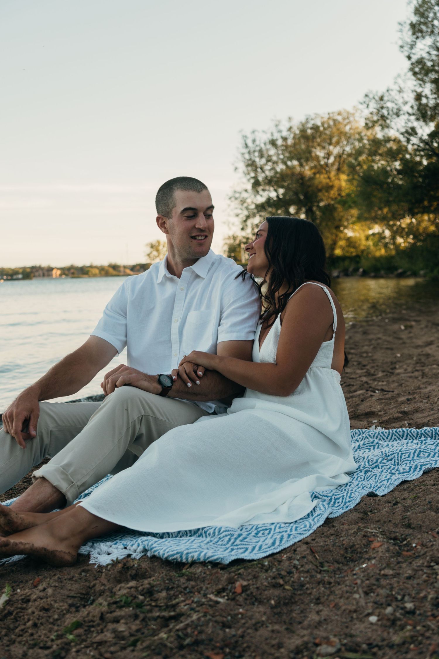 Couple relaxing on blanket by water during sunset engagement session.