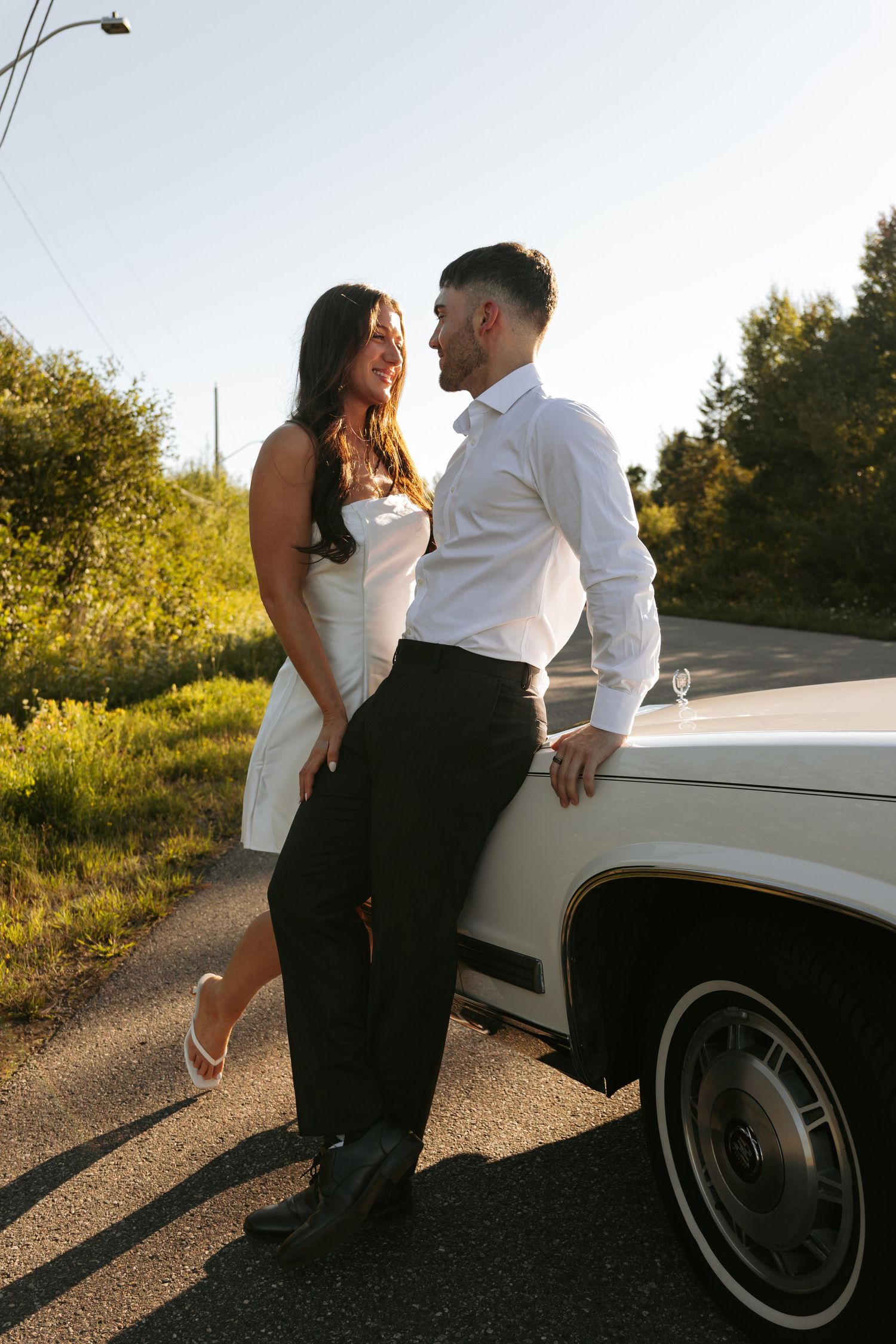 Romantic couple poses against a classic white car during sunset photoshoot.