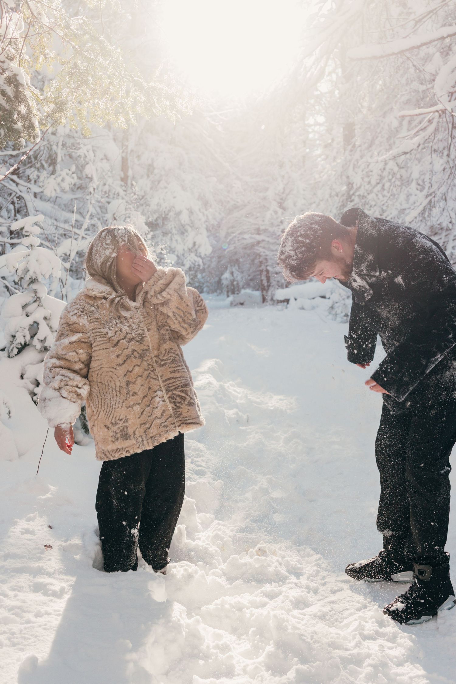 Two people playing and throwing snow at each other in a wintry forest landscape.