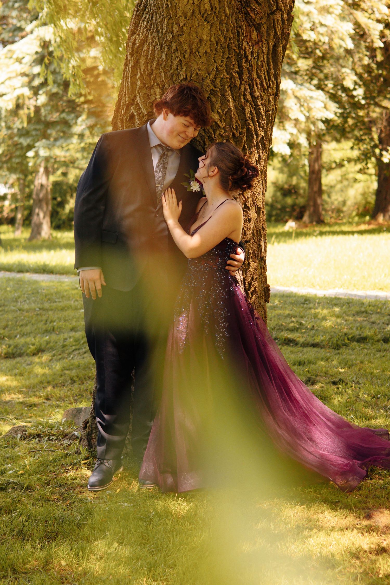 Couple in formal attire sharing a romantic moment by a tree in a sunlit park, woman in purple gown.