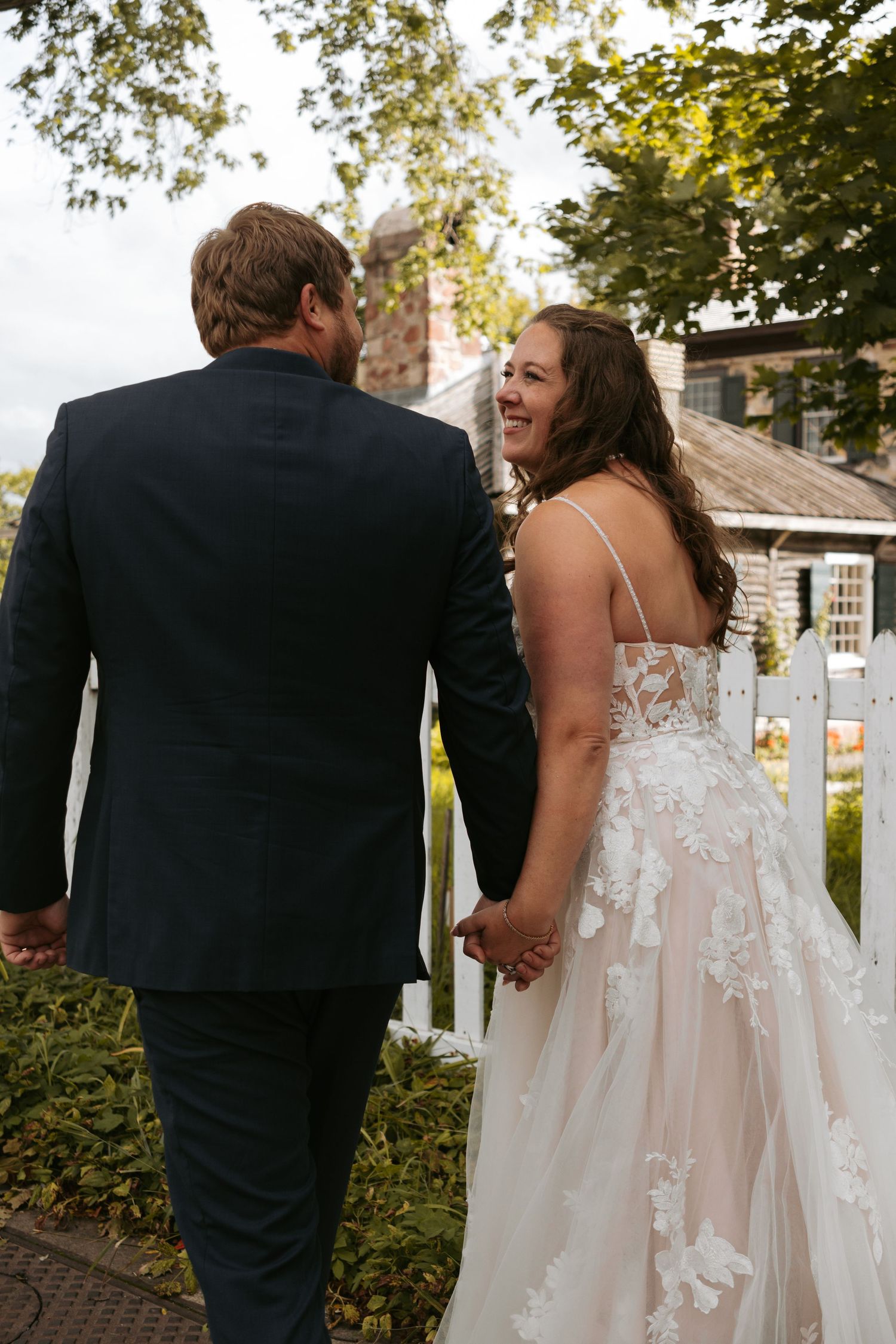Newlyweds walk hand in hand along a garden path with a white picket fence in the background.